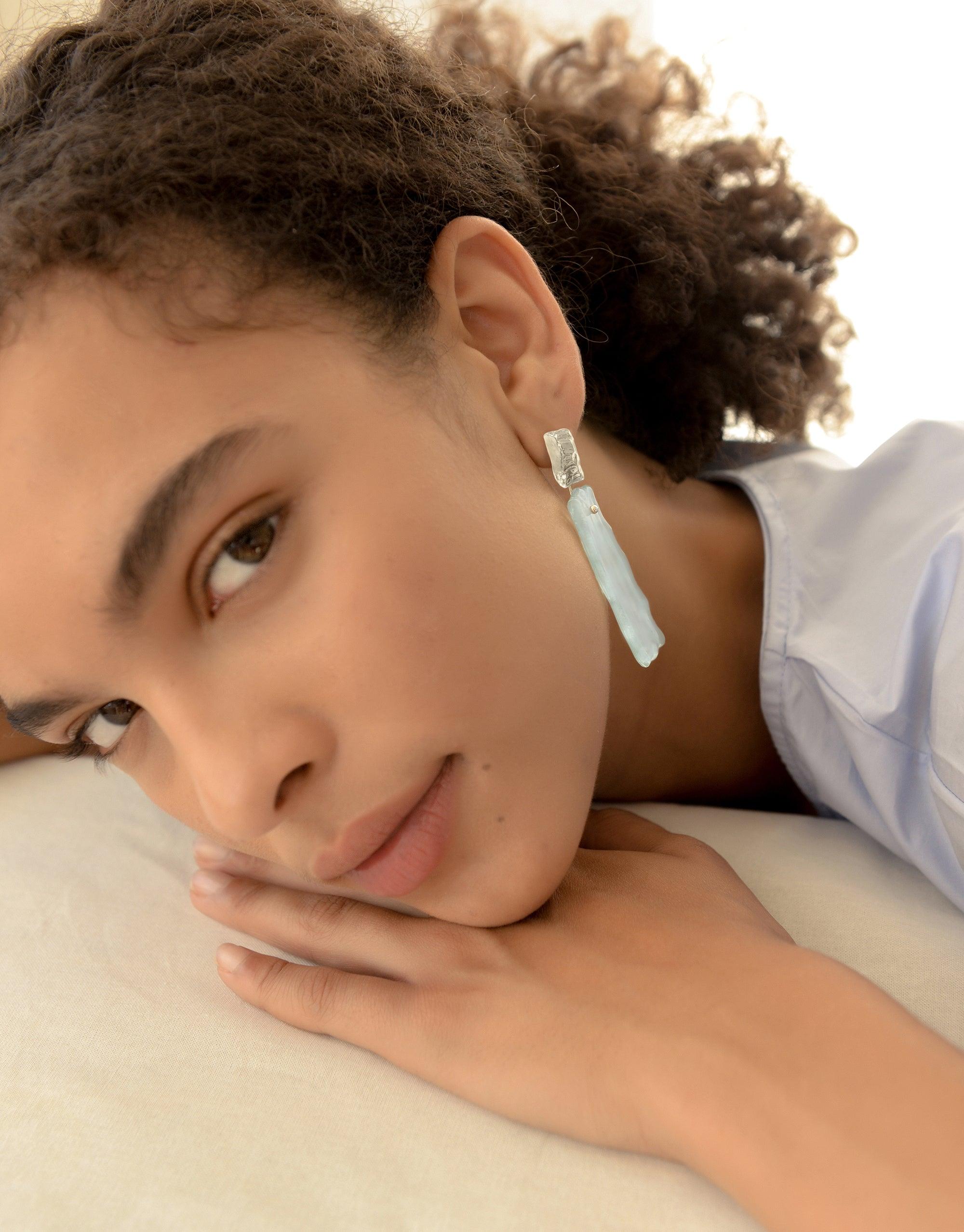 Close-up of elegant Afloat Earrings featuring a pale blue, hand-formed glass pendant hanging from a silver stud. The earrings showcase a unique, organic shape inspired by melting glaciers. Part of the Floating Glacier Collection by CLED.