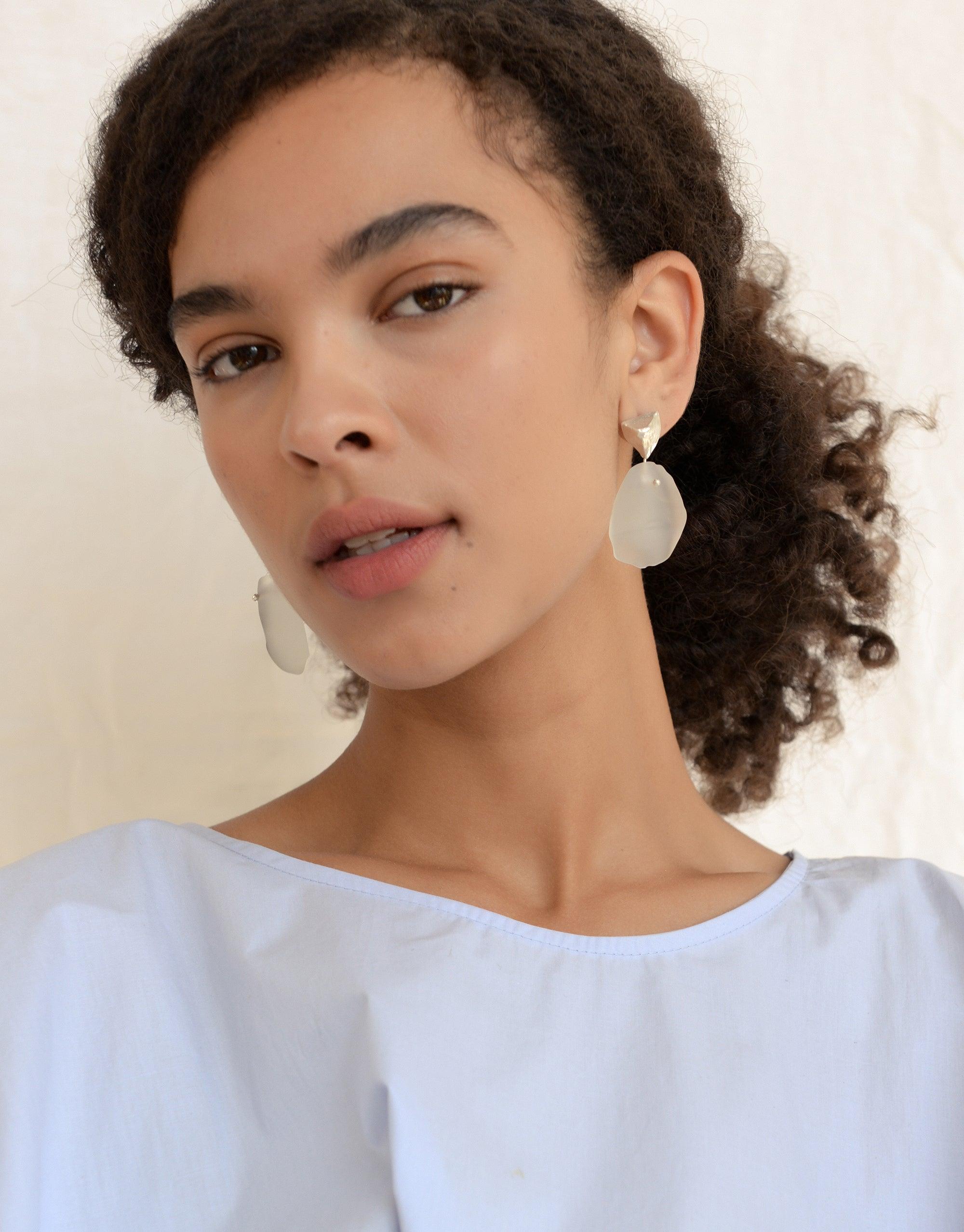 Tip of the Iceberg Earrings: A woman with curly hair showcases sterling silver, matte-finish earrings inspired by icebergs, featuring hand-formed Eco Gems repurposed from discarded glass.