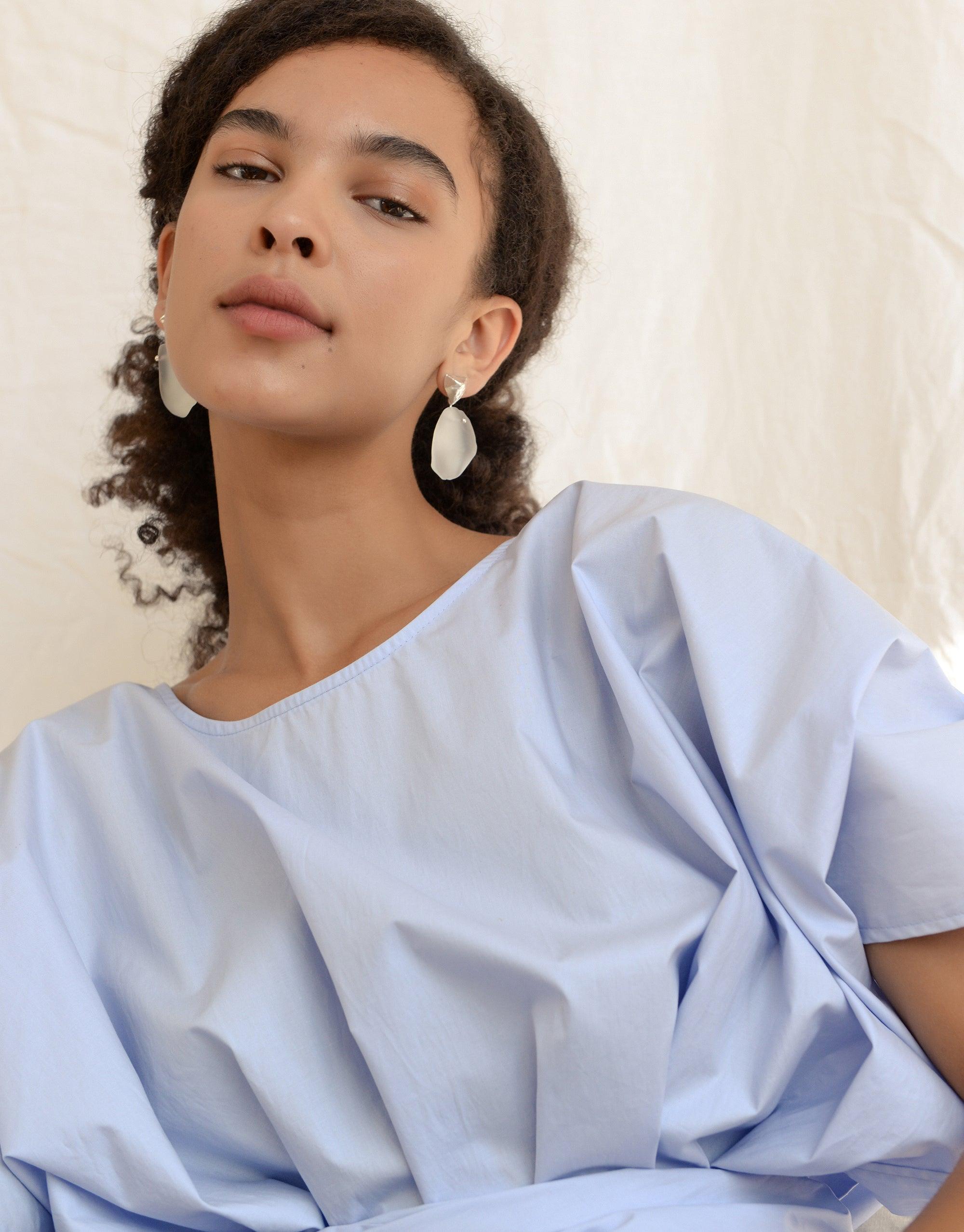 Tip of the Iceberg Earrings: A woman with curly hair wears eco gem drop earrings, handcrafted from repurposed glass, showcasing a matte finish. Part of the Floating Glacier Collection.