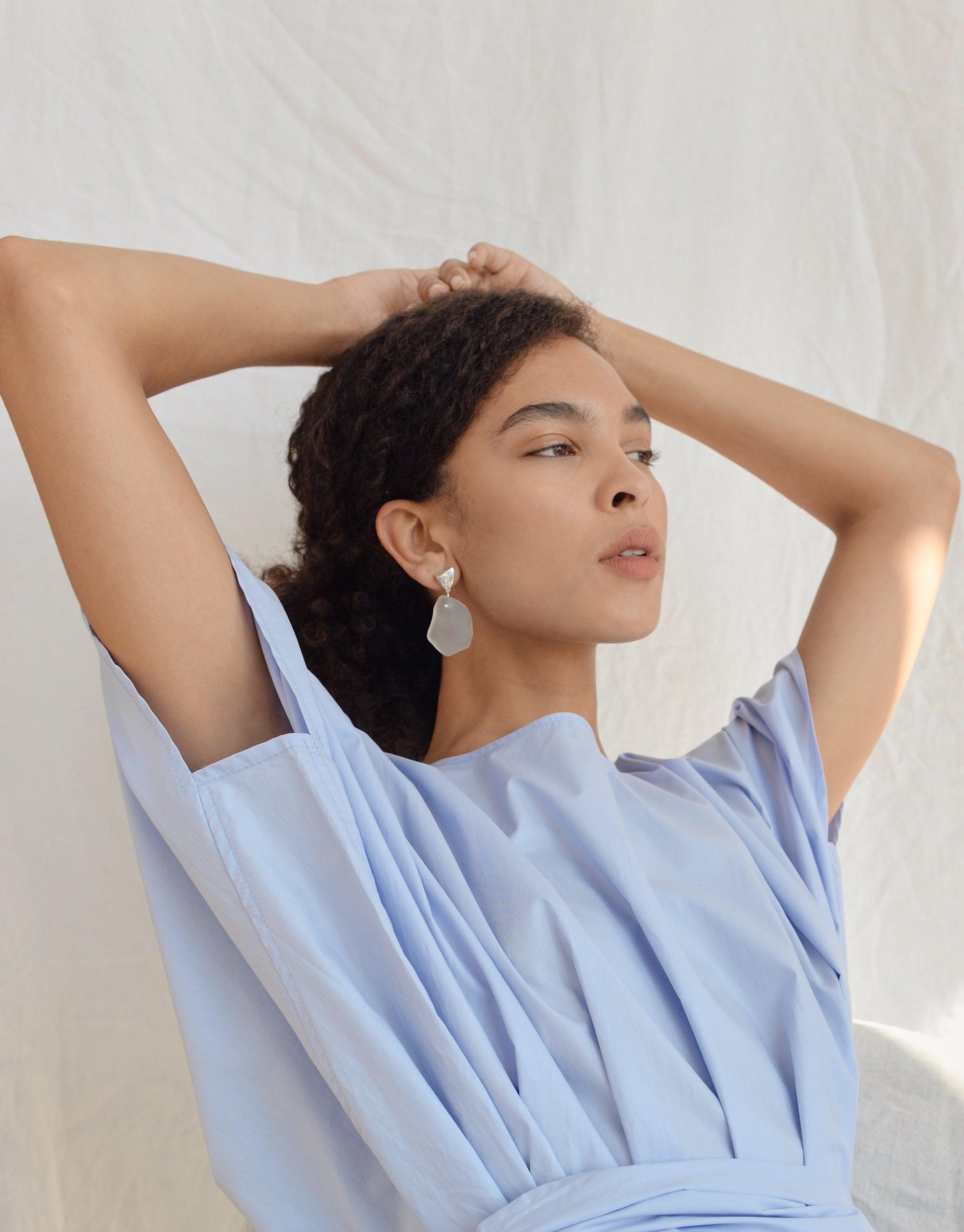 Tip of the Iceberg Earrings: A woman models sterling silver earrings, featuring matte eco gem drops, inspired by Arctic icebergs, with her hand gracefully behind her head.