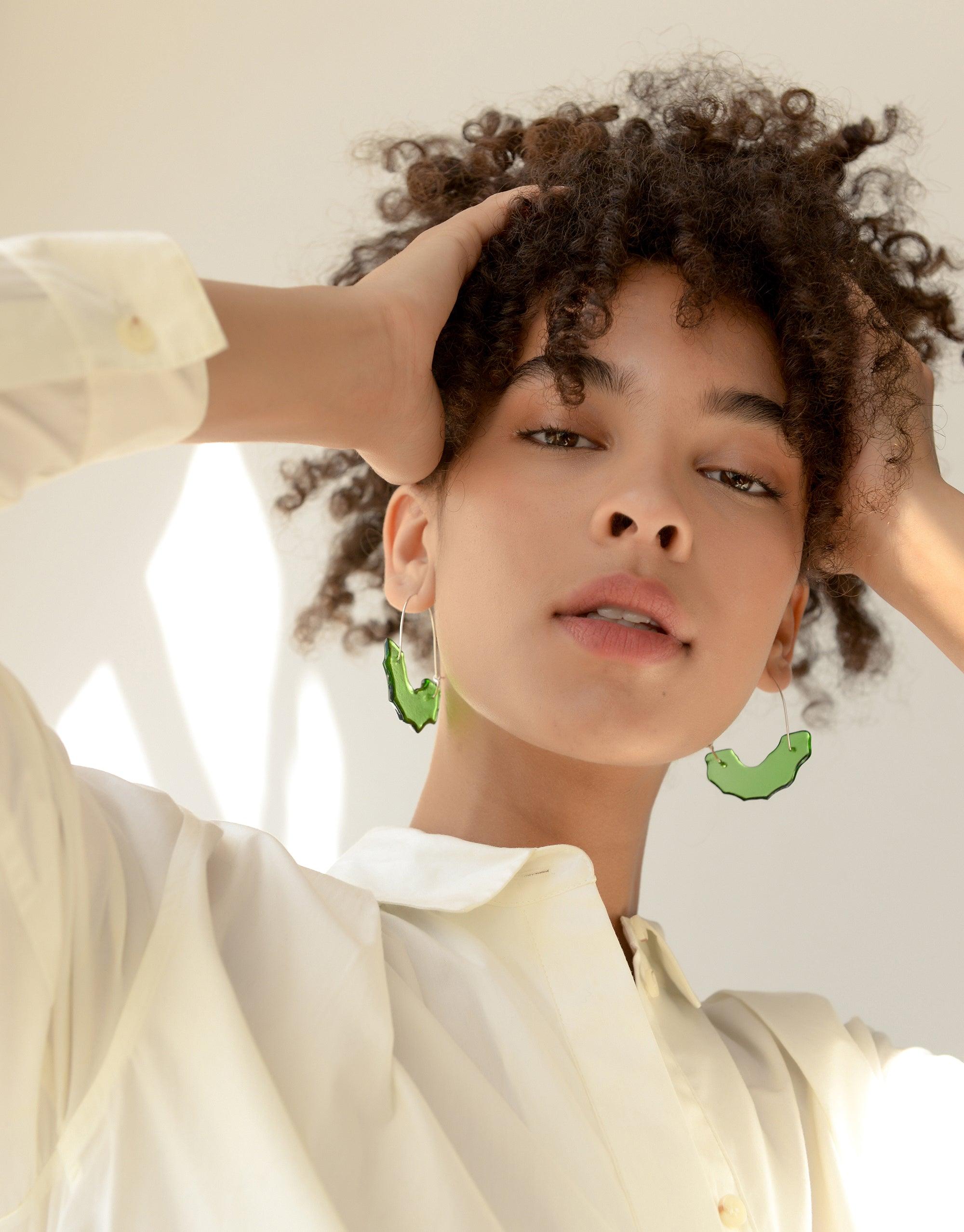 Close-up of model wearing green arch-shaped earrings, showcasing the Polar Arch Hoop Earrings design. Curly hair and natural makeup complement the eco-friendly jewelry. Soft lighting highlights the unique shape and sustainable materials of CLED's Polar Arch Hoop Earrings.
