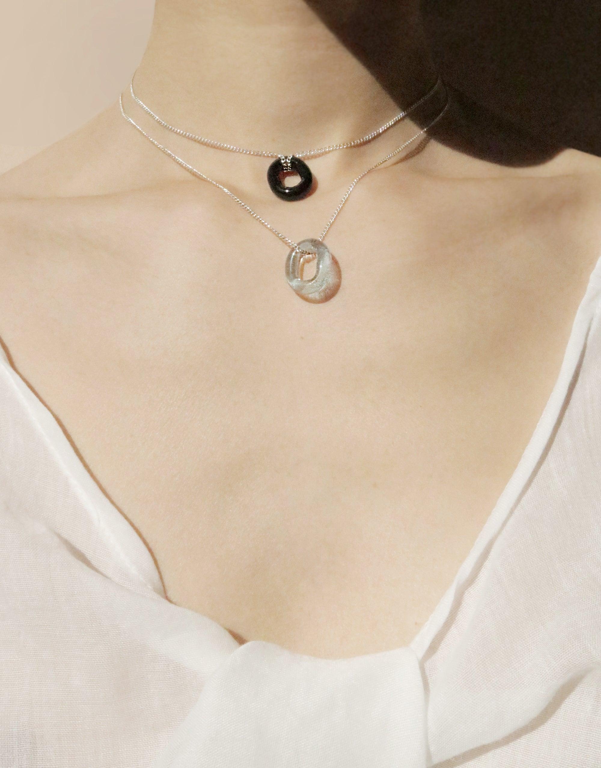 Layered silver necklaces featuring eco-friendly glass pendants with loop designs. The In The Loop Necklace showcases minimalist style with adjustable chain lengths, perfect for versatile wear. Handmade sustainable jewelry by CLED.