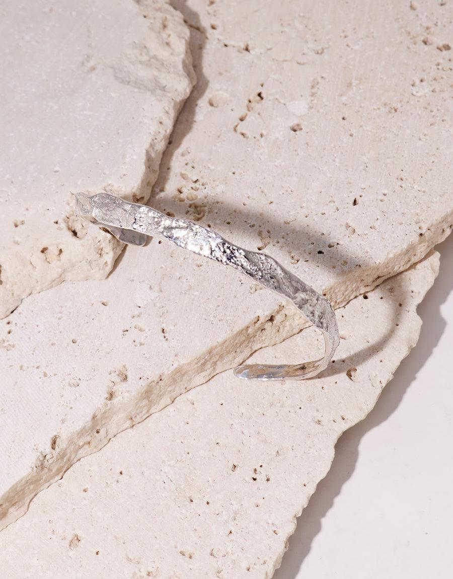 Sterling silver Cliff Bracelet with a textured, organic design resting on a cracked, light-colored stone surface. Handcrafted cuff bracelet showcasing a unique, earthy aesthetic. Versatile and bendable jewelry piece for a custom fit. CLED Cliff Bracelet.