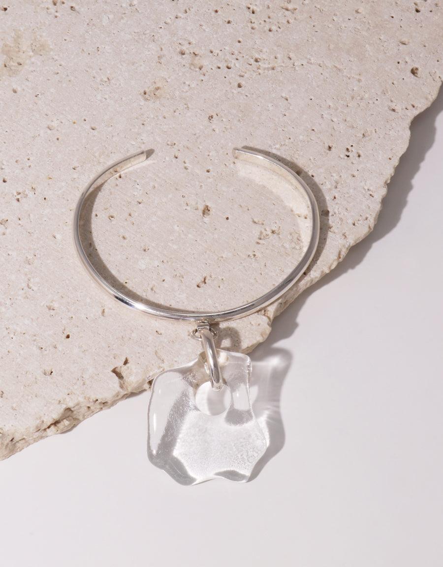 Coastline Drop Bracelet: Sterling silver cuff with unique eco-friendly glass pendant, inspired by seashore shapes. Handcrafted piece resting on textured stone surface, showcasing its elegant design and sustainable materials. CLED