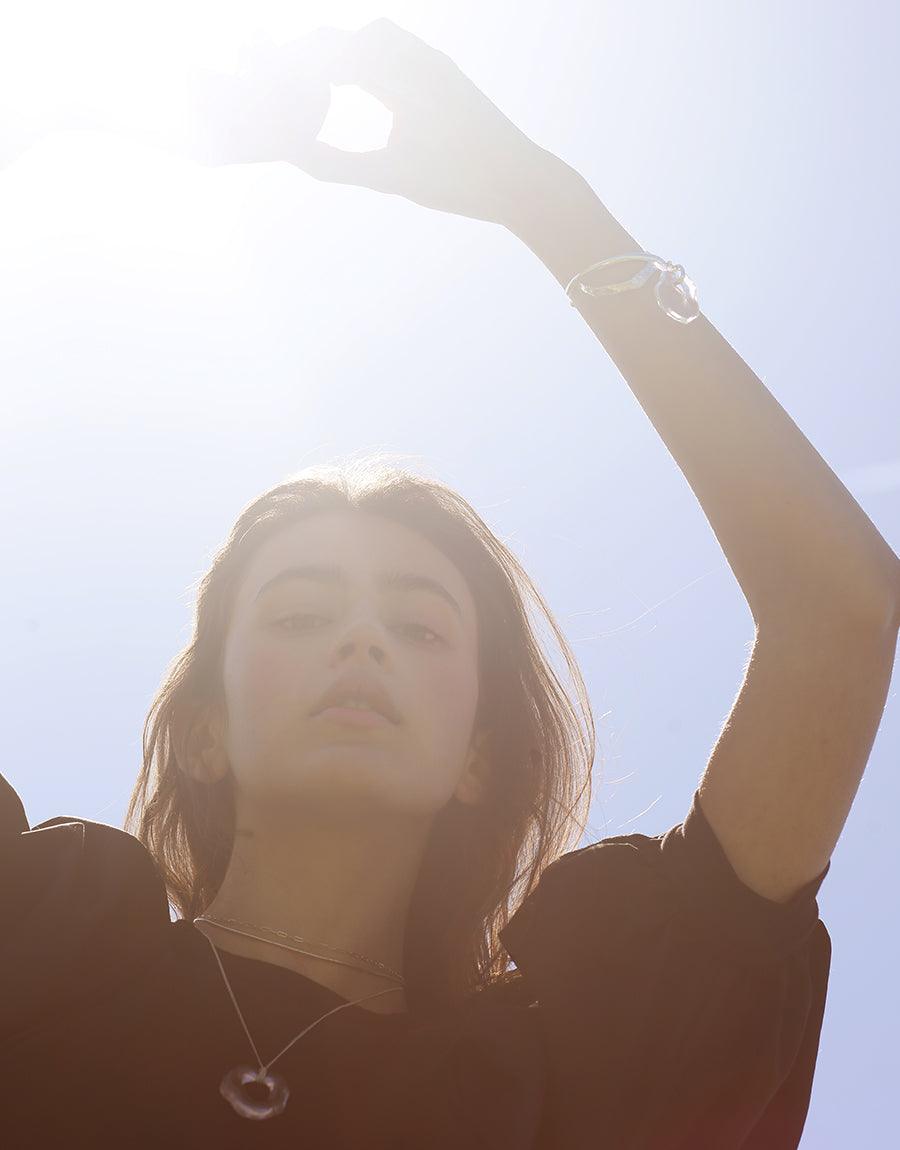 Silhouette of a person with raised arm against bright sunlight, wearing a cuff bracelet that catches the light, showcasing the elegant design of the Coastline Drop Bracelet. Handcrafted jewelry by CLED.