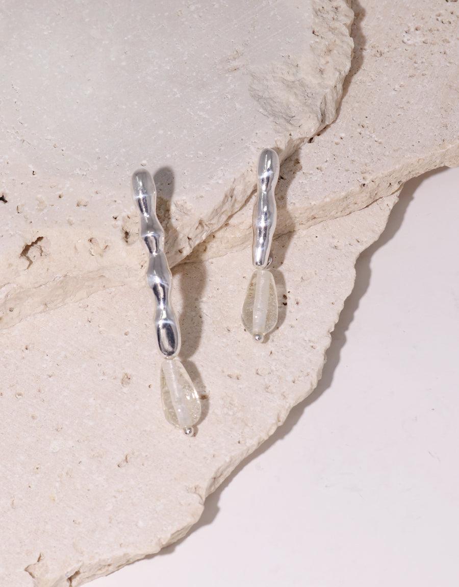 Water Earrings, handcrafted in sterling silver with unique eco gems repurposed from glass bottles, displayed on a rock. Each asymmetrical piece reflects CLED's commitment to mindful conservation.