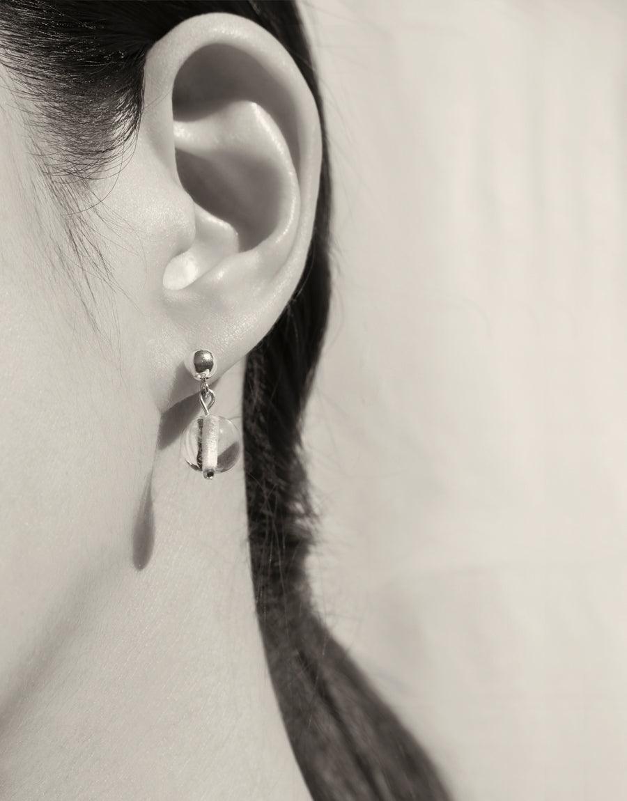 Close-up of an ear wearing a delicate drop earring with a small, transparent bead. The minimal design showcases the elegant simplicity of the Minimal Ball Earrings | Small. Handcrafted from sterling silver and eco-friendly glass by CLED.
