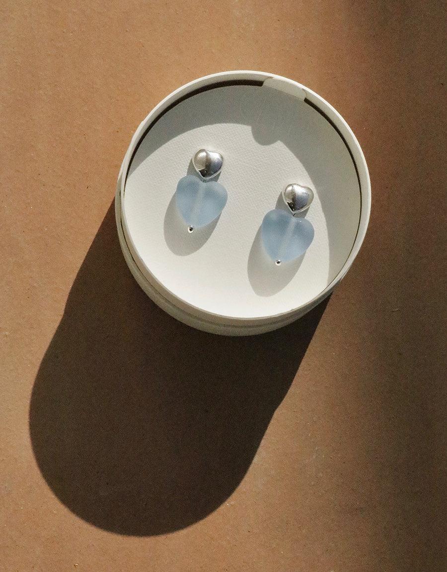 Elegant Love Earrings with Large Heart Drop featuring matte blue jay eco-friendly glass hearts in a circular white display. Sterling silver studs complement the delicate design. Handcrafted sustainable jewelry by CLED.