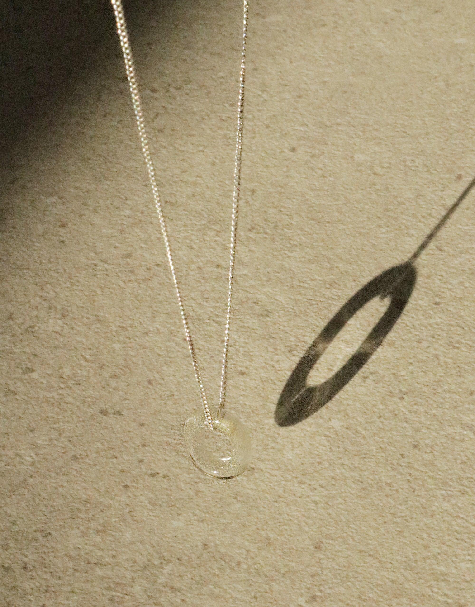 Delicate silver chain necklace with transparent eco-glass loop pendant, casting oval shadow on beige surface. Minimalist design showcases sustainable elegance in "In The Loop Necklace" by CLED.