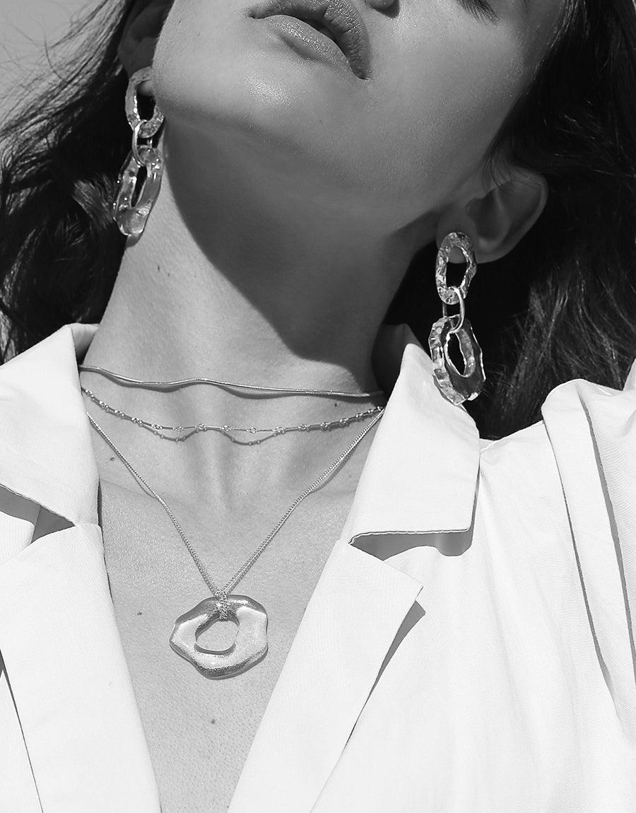 Black and white image showcasing the High Tide Necklace, featuring an organically shaped pendant on a delicate chain. Model wearing a white blazer, paired with statement earrings. Emphasizes the necklace's versatile and elegant design, perfect for layering. Handcrafted jewelry by CLED.
