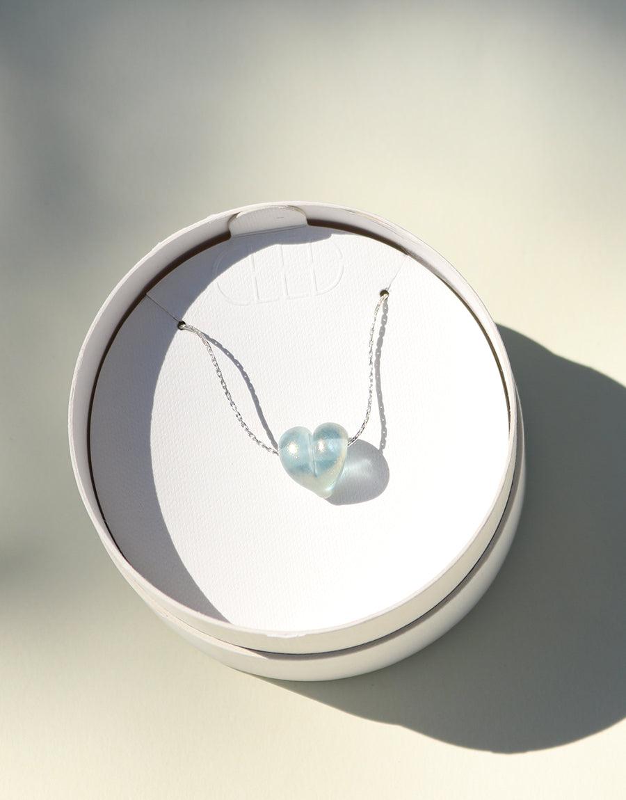 Love Necklace featuring a delicate heart-shaped pendant made from repurposed glass, displayed on a white circular platform. The necklace's sterling silver chain shimmers, showcasing the sustainable and handcrafted nature of this elegant piece by CLED.