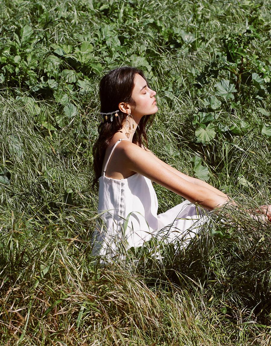 Woman in white dress sitting in lush green field, eyes closed, enjoying nature. Floral Barrette by CLED visible in her hair, showcasing the product's elegance in a natural setting.