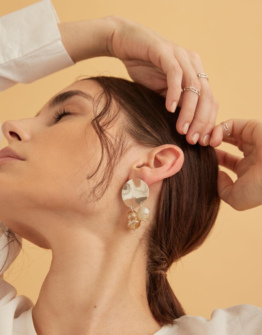 Elegant model showcasing delicate silver jewelry, including Collapsible Chain Ring | C, earrings, and multiple rings on fingers. Hands gracefully positioned near face, emphasizing the versatile and stackable nature of the jewelry. Soft lighting and warm background highlight the handcrafted, Los Angeles-made CLED pieces.
