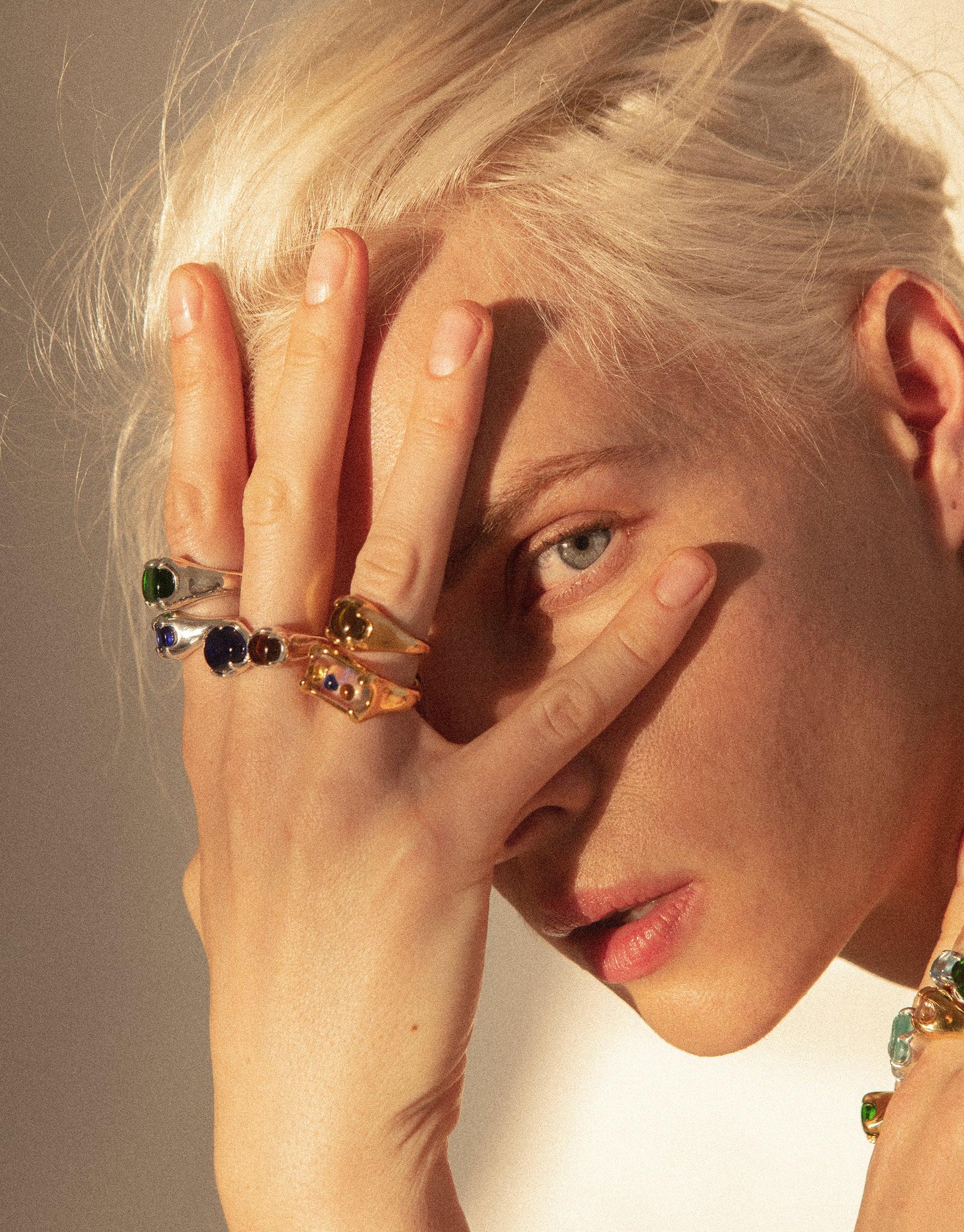 Close-up of a hand wearing multiple colorful rings, including the Prism Ring in Gold Plated, showcasing eco gems upcycled from discarded glass. The hand partially covers a face, highlighting the jewelry's vibrant colors and unique design. Prism Ring | Gold Plated by CLED.