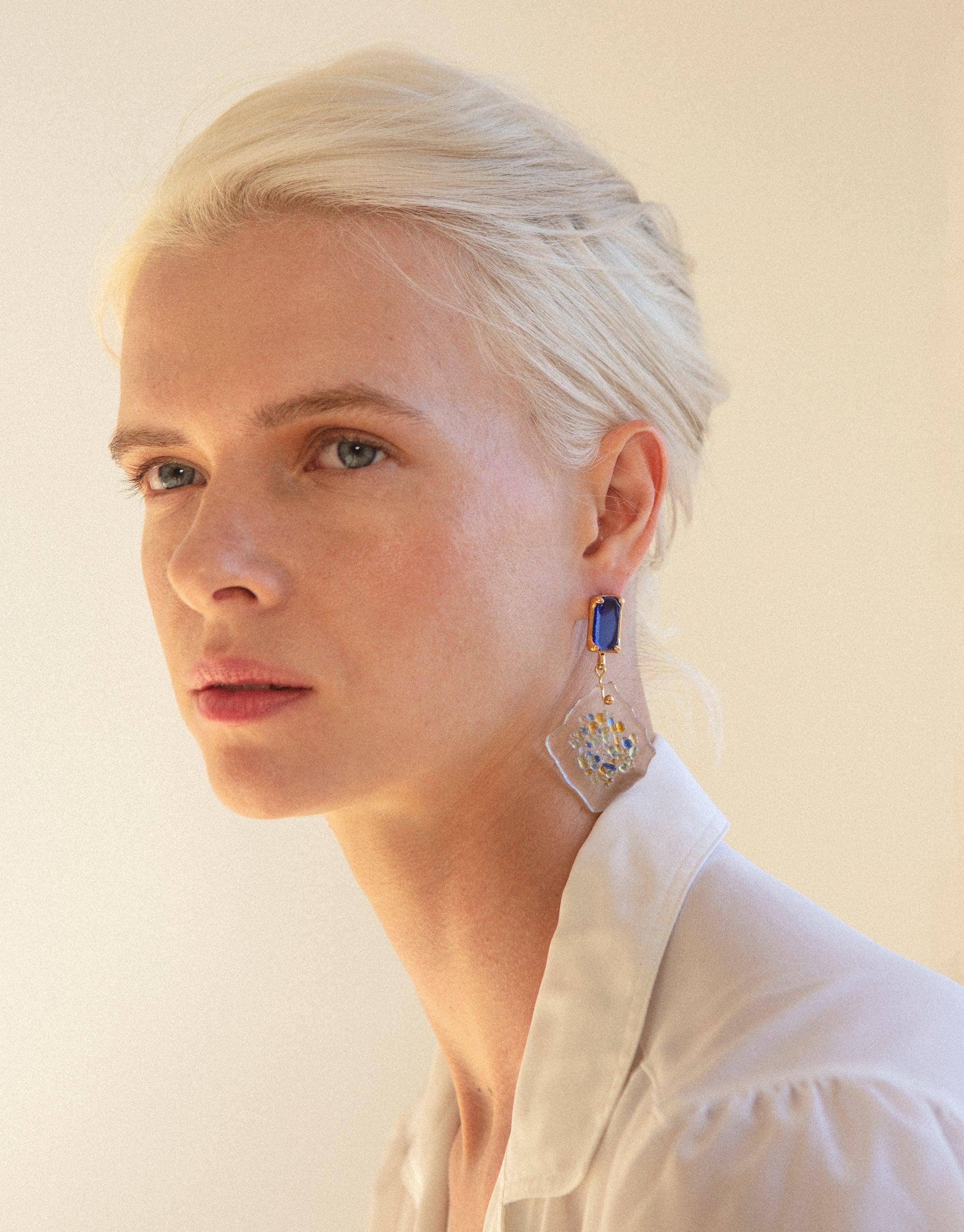 Model wearing Spectral Prism Earrings featuring blue eco gem studs and white diamond-shaped pendants with colorful glass fragments, showcasing sustainable jewelry design inspired by prismatic light refraction. Handmade, upcycled glass earrings by CLED.