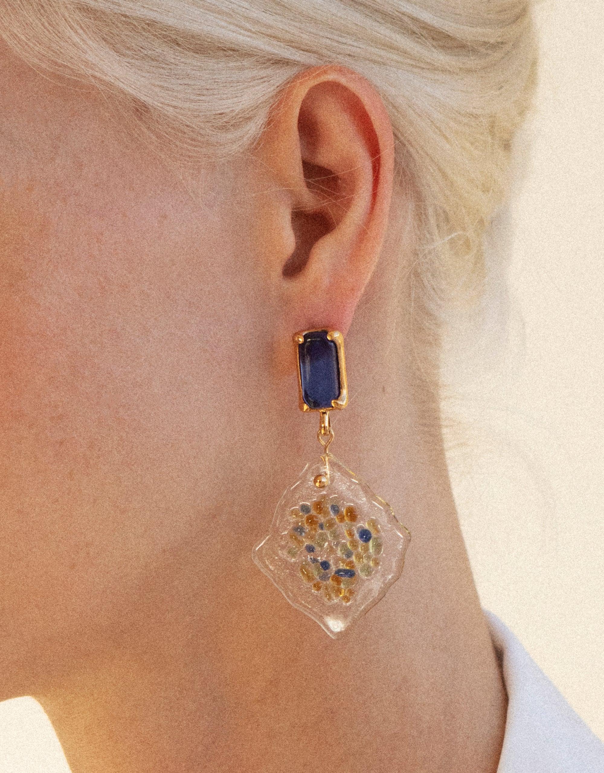 Close-up image of a model wearing Spectral Prism Earrings featuring blue eco gem studs and white diamond-shaped pendants with colorful glass fragments, showcasing sustainable jewelry design inspired by prismatic light refraction. Handmade, upcycled glass earrings by CLED.