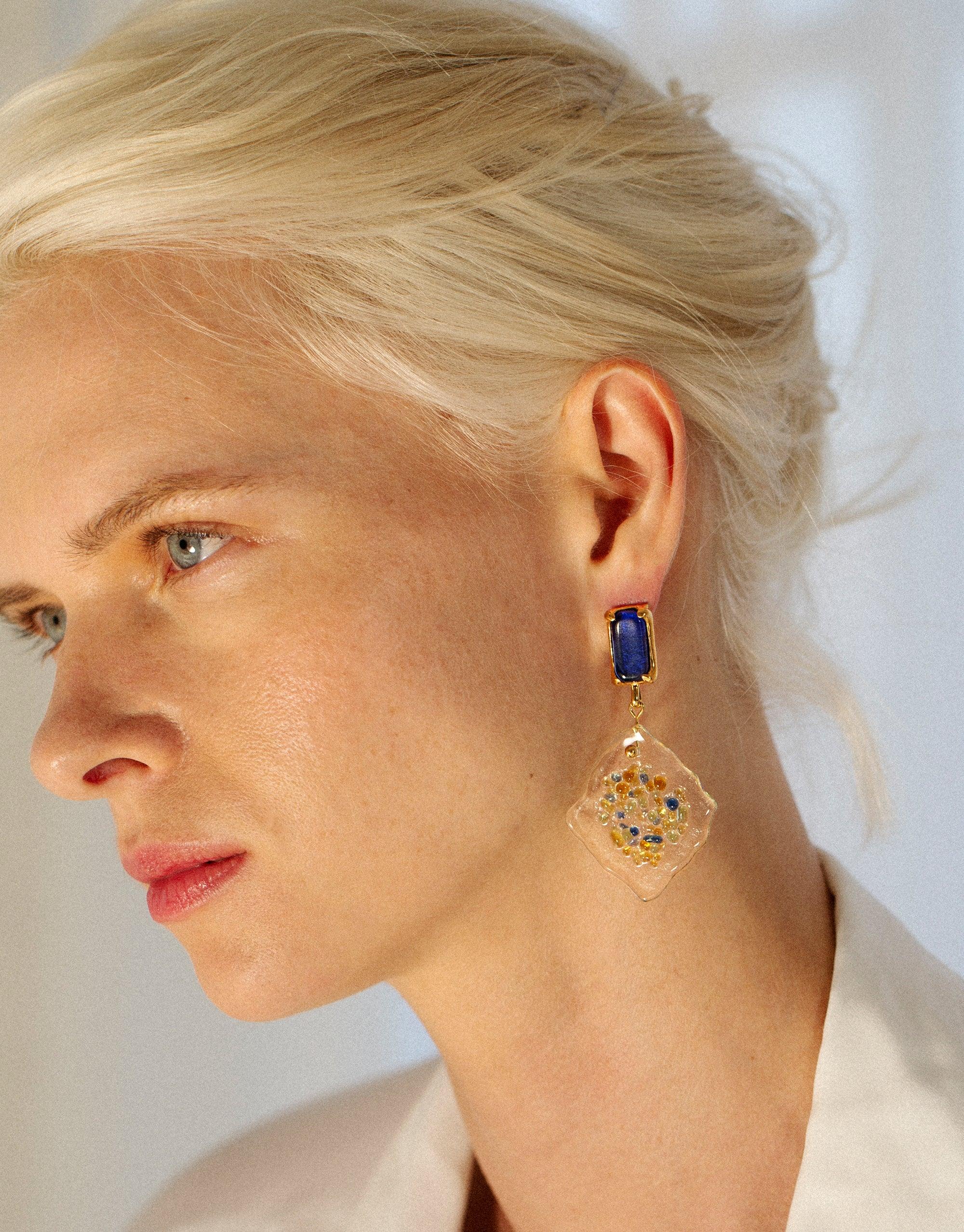 Model wearing Spectral Prism Earrings featuring blue eco gem studs and white diamond-shaped pendants with colorful glass fragments, showcasing sustainable jewelry design inspired by prismatic light refraction. Handmade, upcycled glass earrings by CLED.