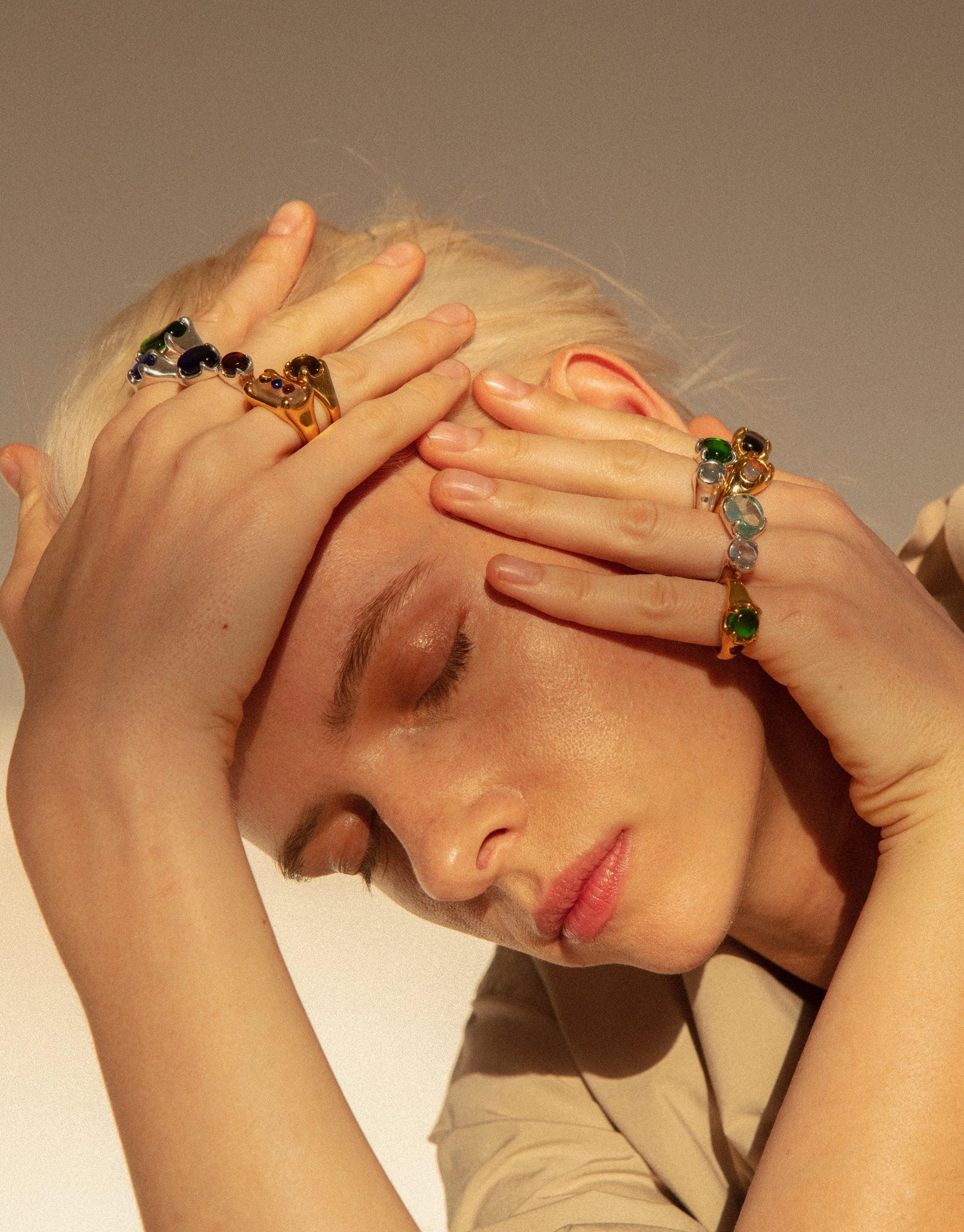 Close-up of hands adorned with colorful Beam Rings, showcasing eco gems in various hues. Model's face partially visible, emphasizing the vibrant, prism-like effect of the jewelry. Handcrafted sustainable accessories by CLED.
