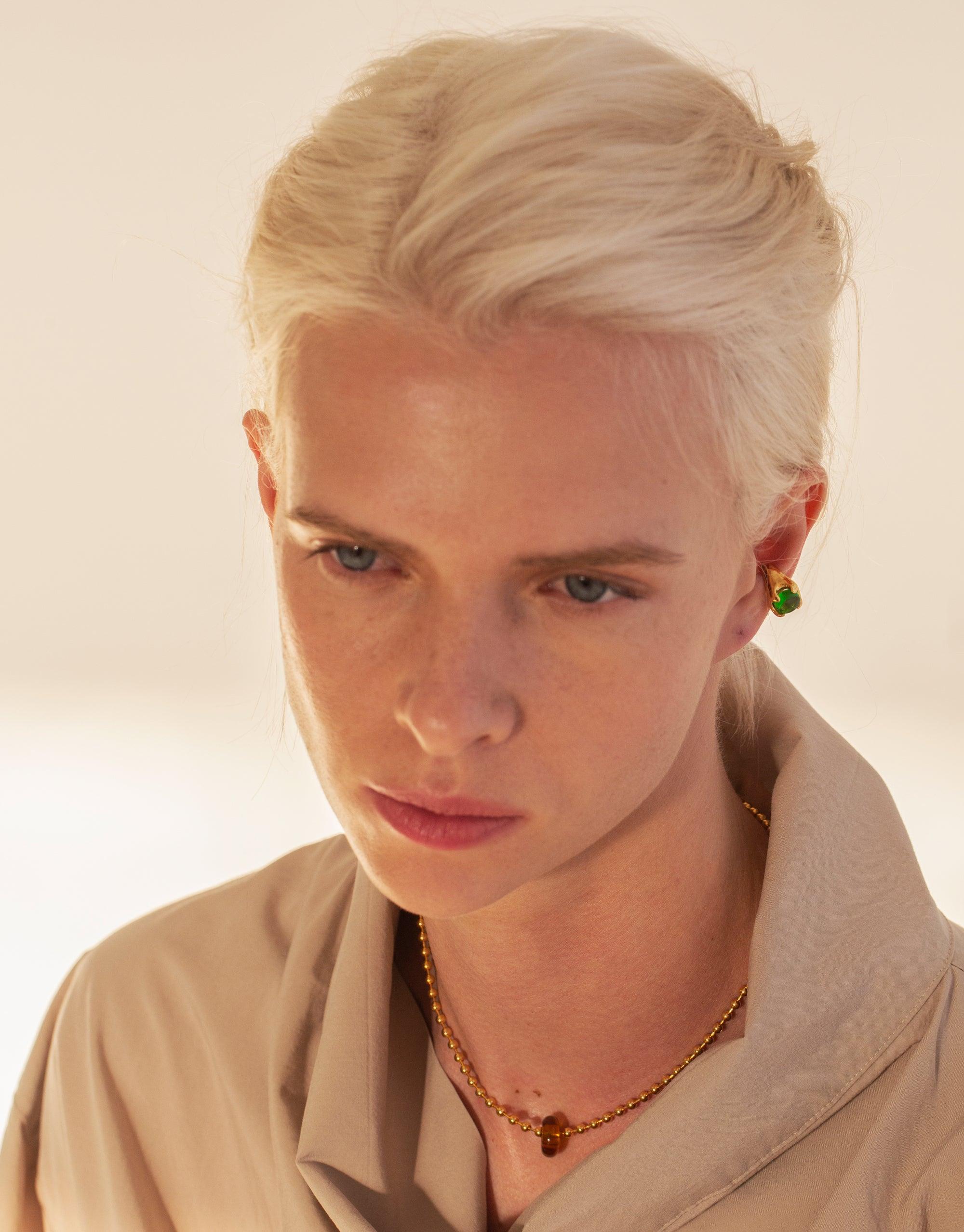 Close-up portrait of a model wearing a delicate gold necklace featuring a small, spherical pendant, showcasing the Donut Ball Necklace. The necklace rests elegantly on the collarbone, highlighting its minimalist design and craftsmanship by CLED.