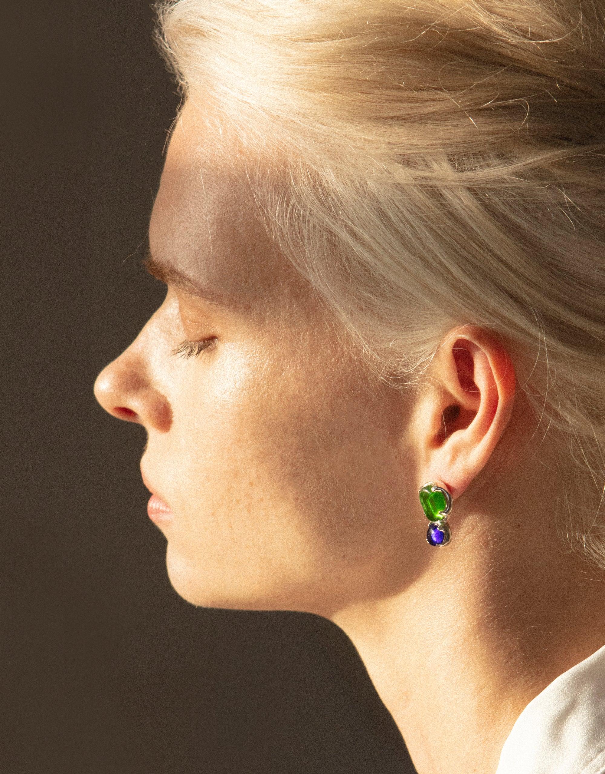 Profile view of a person wearing Duo Beam Earrings, showcasing vibrant green and blue eco gems repurposed from glass. The earrings catch light, creating a prism-like effect against blonde hair. Elegant and sustainable jewelry by CLED.