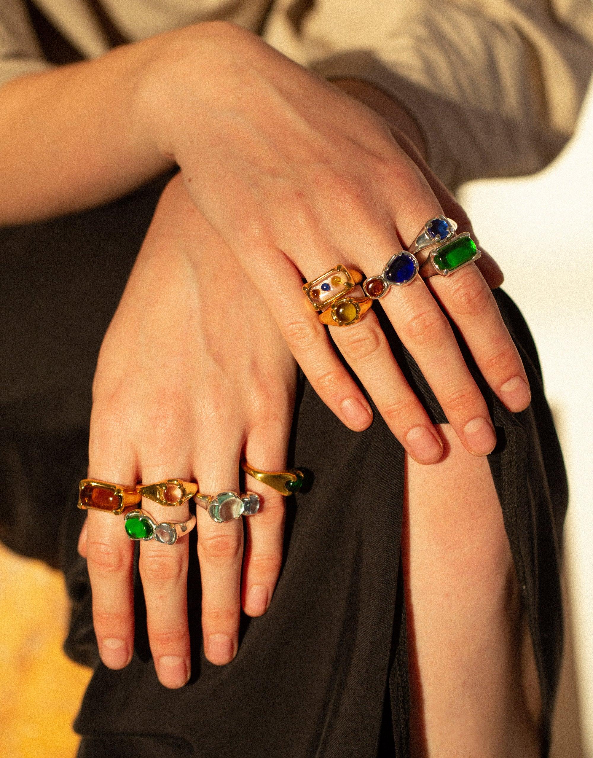 Hands adorned with colorful eco-friendly rings, showcasing the Prism Ring Gold Plated collection. Vibrant upcycled glass gems in various hues reflect light, reminiscent of a prism's spectrum. Sustainable jewelry design by CLED featuring the Prism Ring | Gold Plated.