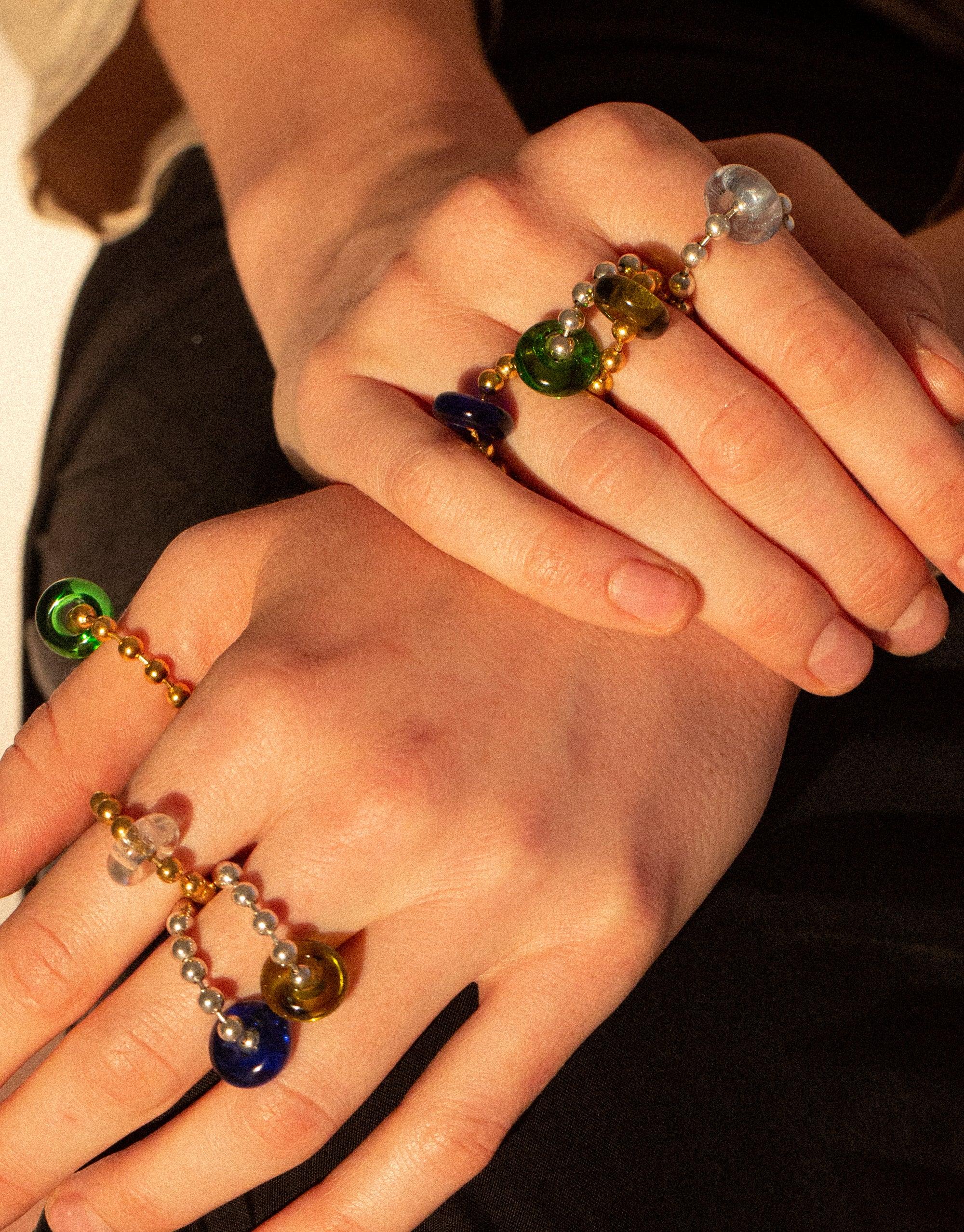 Close-up of hands wearing colorful Donut Ball Rings, featuring vibrant eco gems in various hues reminiscent of prismatic light. Each ring showcases uniquely shaped glass beads, highlighting the sustainable and artistic craftsmanship of the CLED Donut Ball Ring collection.