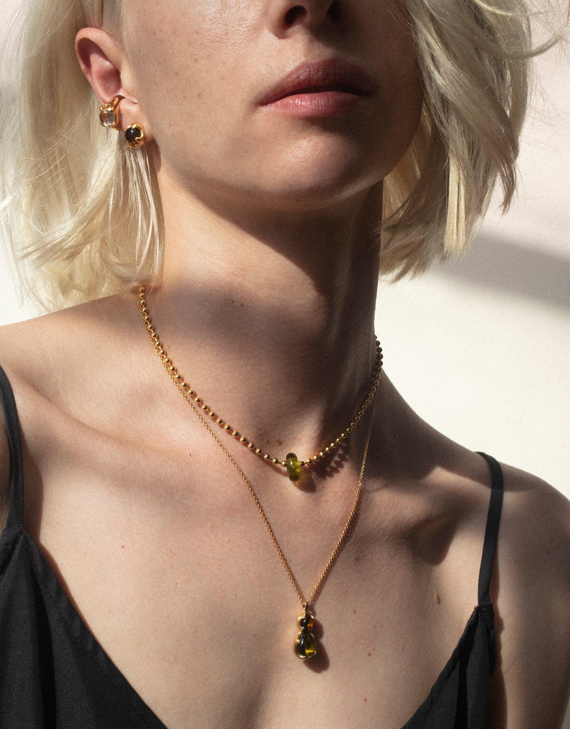 Close-up of a woman wearing layered gold necklaces featuring colorful eco gems, showcasing the Donut Ball Necklace. Sunlight highlights the prism-like effect of the jewelry, embodying the Rays of Light collection by CLED.