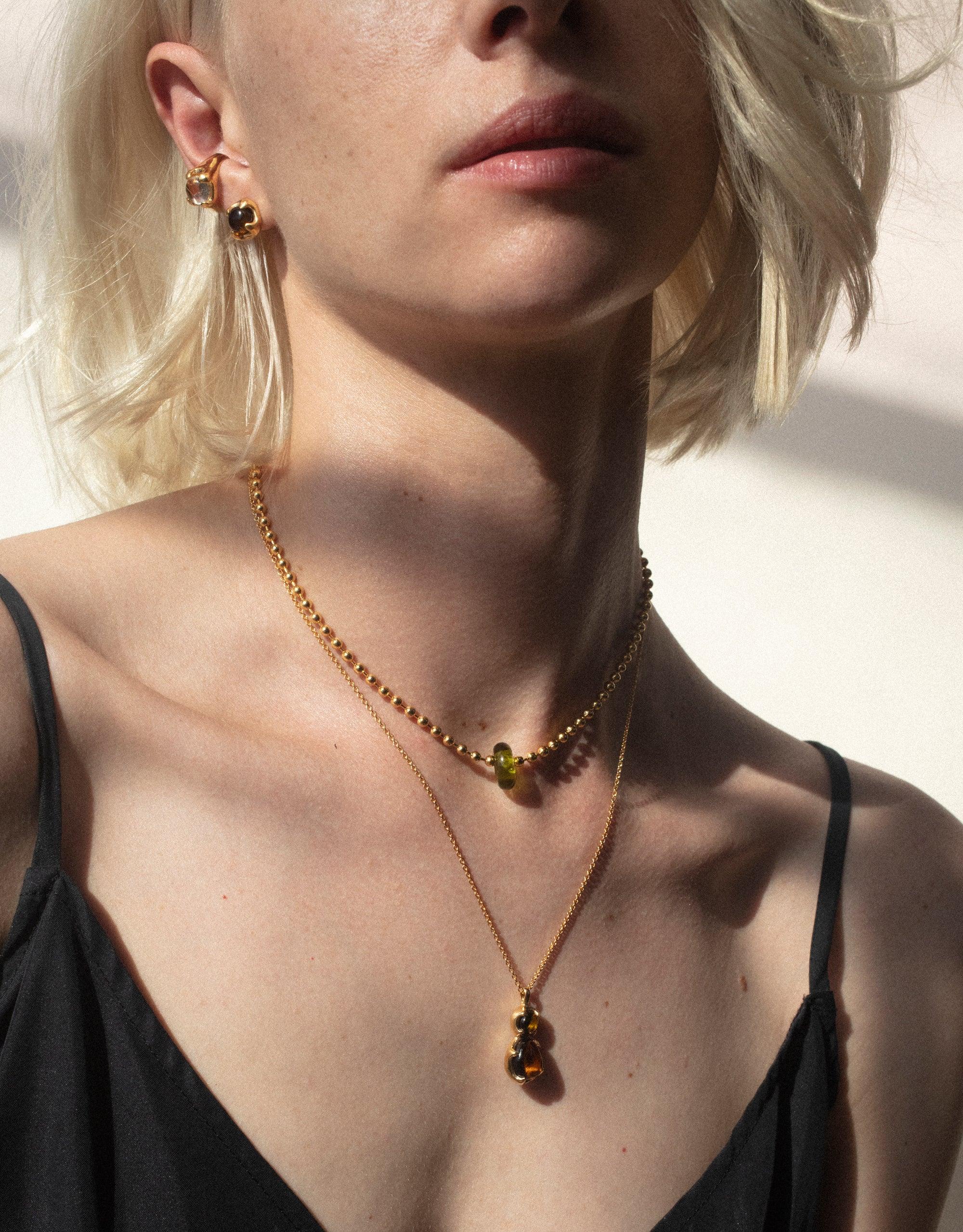 Close-up of a person wearing gold jewelry, including a delicate necklace and ear cuff. The Beam Ear Cuff by CLED showcases eco-friendly gems repurposed from glass, capturing the essence of prism spectral colors in a sustainable design.