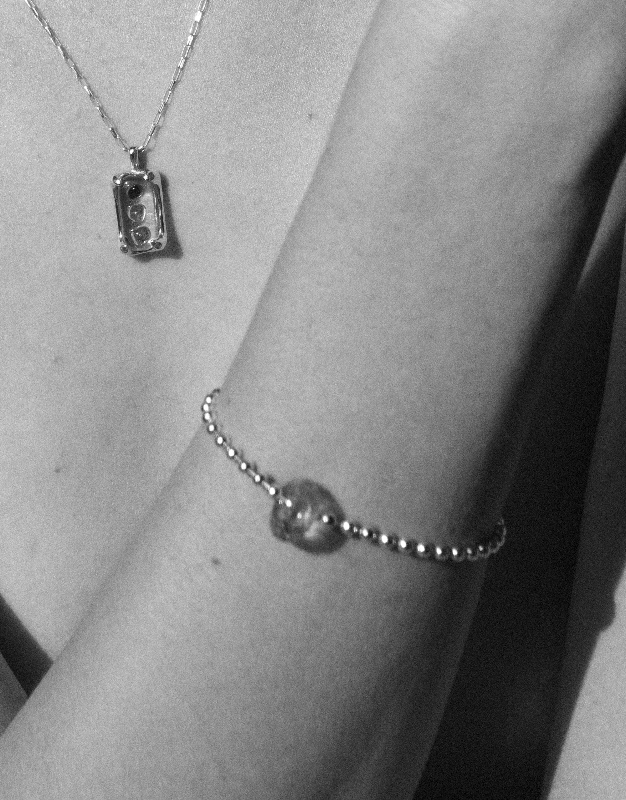 Black and white image showcasing Donut Ball Bracelet on a wrist, featuring a delicate chain with a spherical eco gem centerpiece. A pendant necklace with rectangular charm is visible in the background, highlighting CLED's sustainable jewelry design.