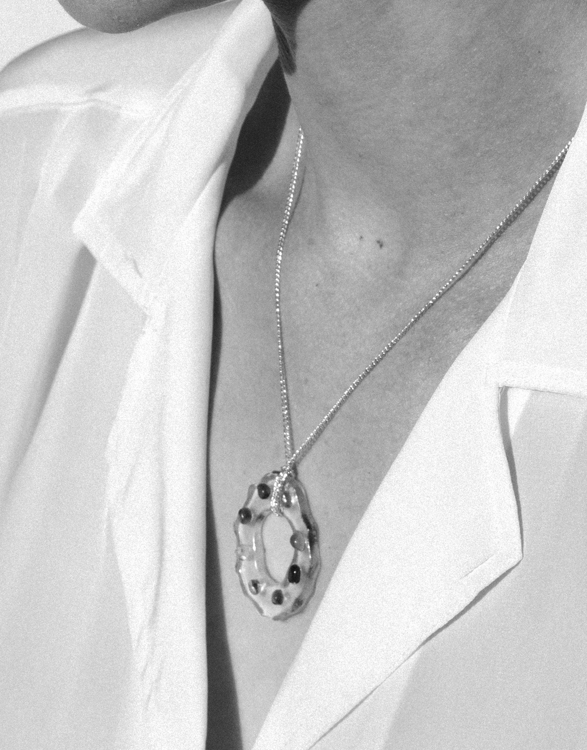High Tide Necklace in Spectrum featuring a delicate silver chain with a unique circular pendant. The pendant showcases a transparent, wave-like design with embedded colorful elements, creating a prism effect. Worn against a white shirt, the necklace exudes elegance and eco-conscious style. CLED