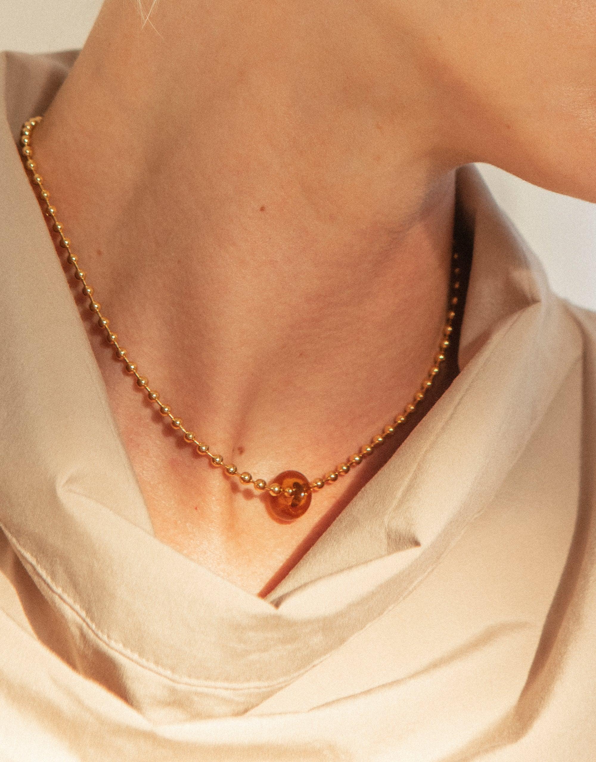 Elegant Donut Ball Necklace featuring a delicate gold chain with a unique amber-colored eco gem pendant, showcasing sustainable luxury against a soft white fabric backdrop. Handcrafted from upcycled glass, this prism-inspired piece reflects CLED's commitment to eco-friendly jewelry design.