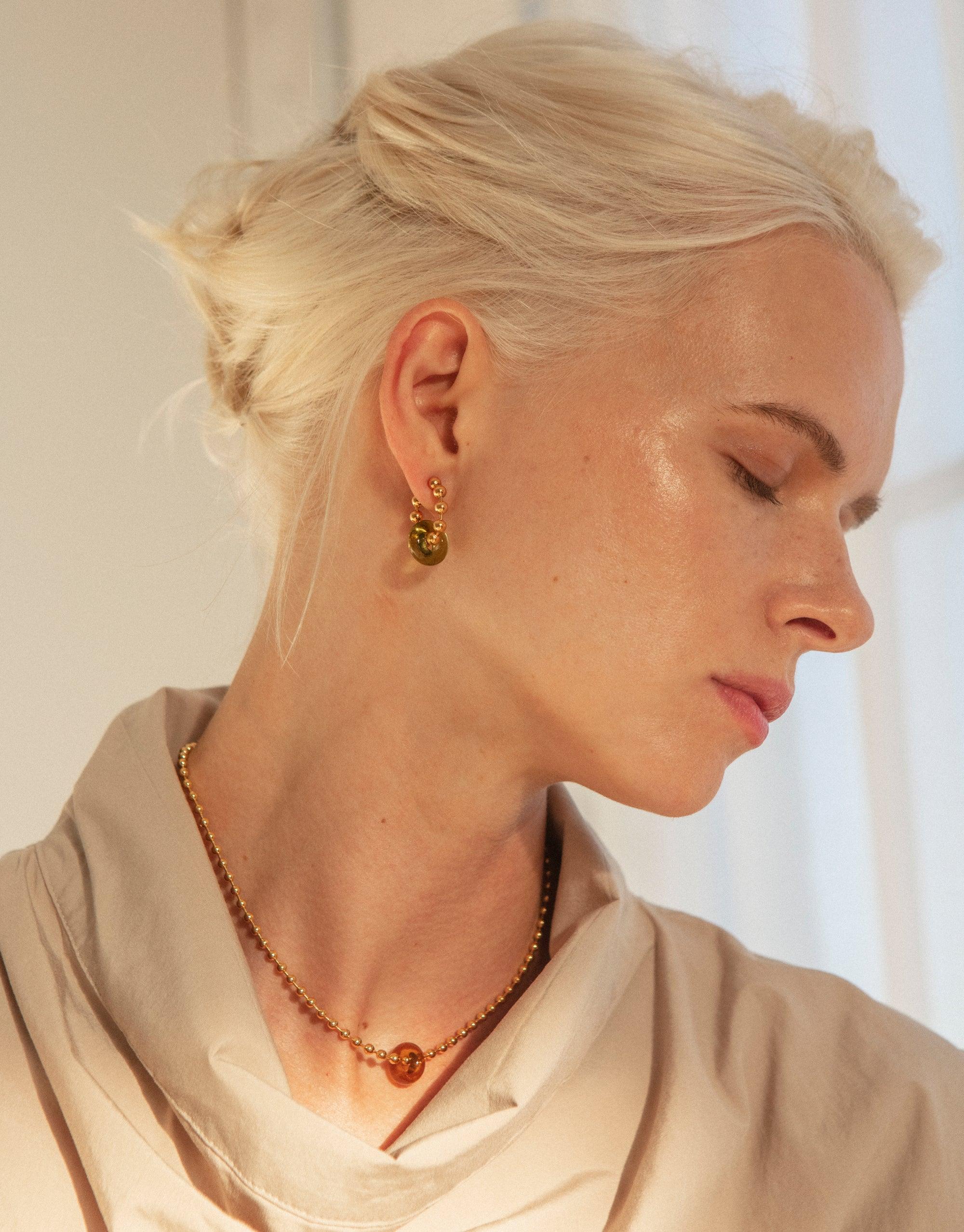 Profile view of a person wearing a gold Donut Ball Necklace with a matching earring, showcasing the elegant design and eco-friendly materials. The necklace features a unique spherical pendant, reflecting CLED's commitment to sustainable jewelry. Donut Ball Necklace by CLED.
