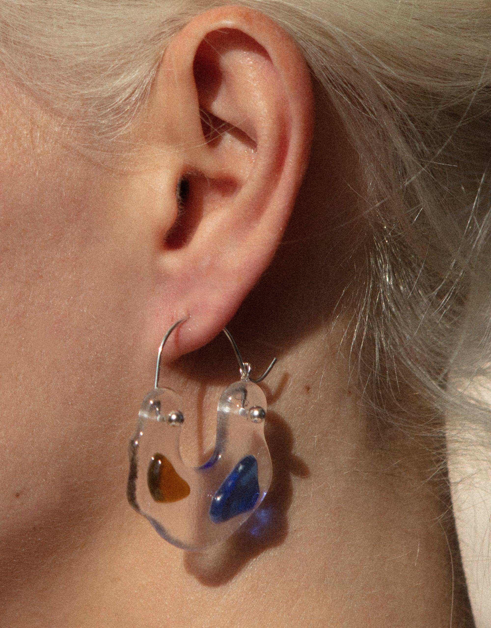 Close-up of Ray Hoop Earrings worn on an ear, showcasing transparent glass hoops with colorful eco gems in amber and blue. Handcrafted sterling silver hooks. Sustainable jewelry by CLED.