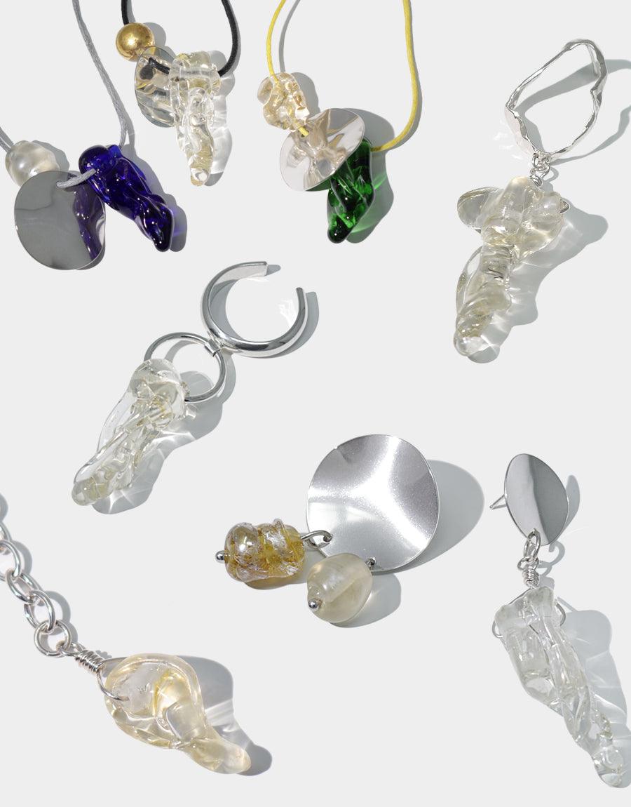 A collection of eco-conscious jewelry pieces, including earrings of various shapes, a silver ring with a clear ball, and a clear organic shaped ring, displayed on a white surface.