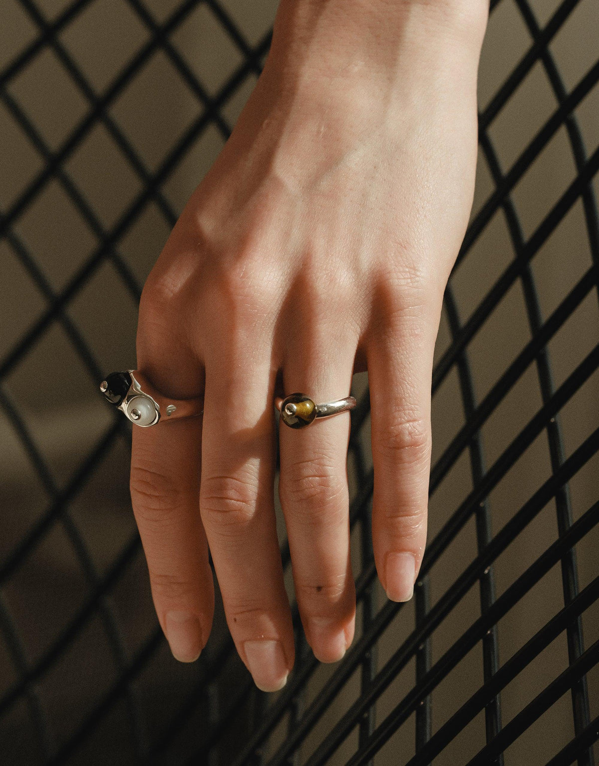 Hand wearing two unique rings with curved designs, showcasing the Pea Curve Ring | Large. The rings feature organic shapes inspired by legume patterns, reflecting nature's creativity and growth. Elegant jewelry against a patterned background. CLED