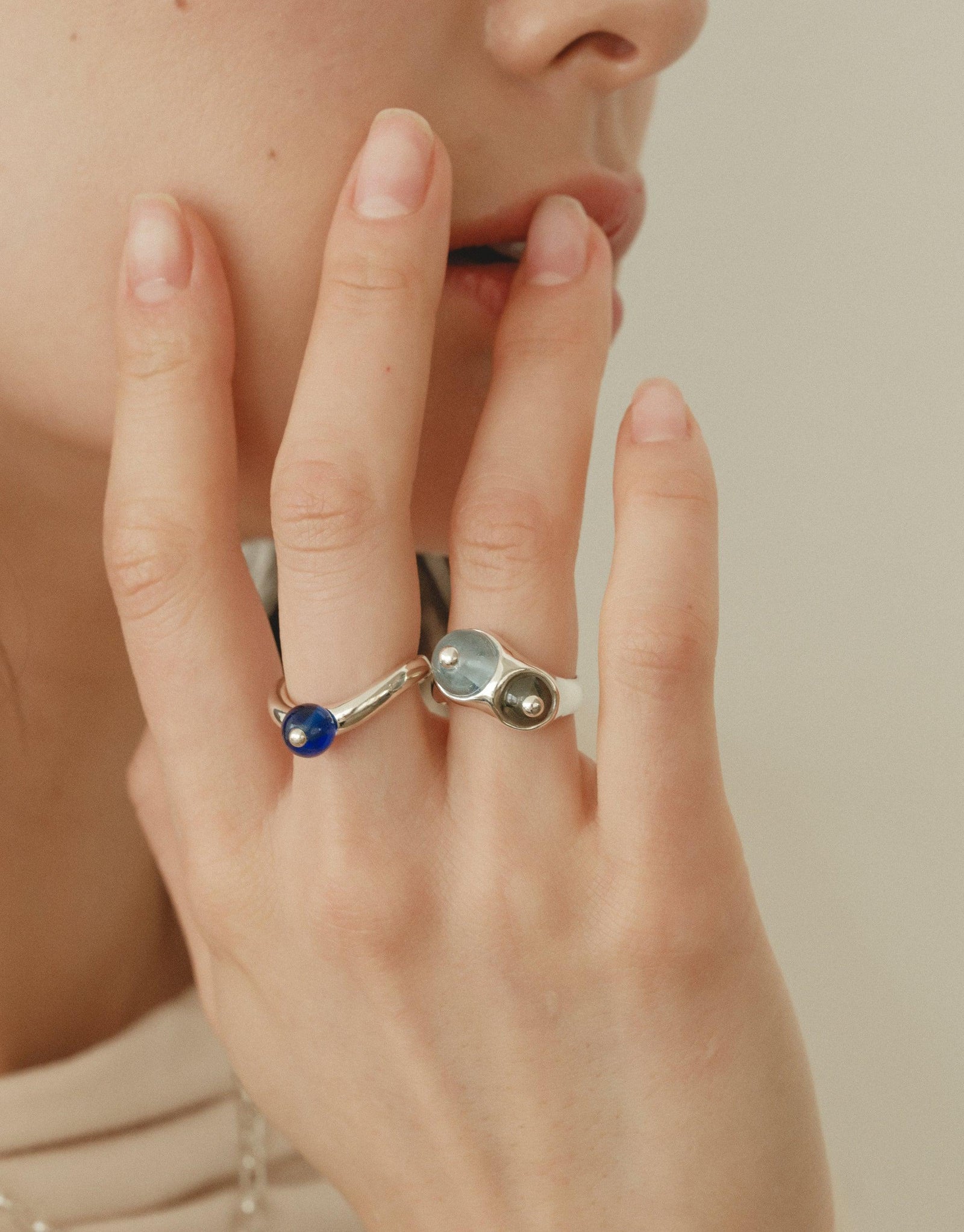 Close-up of a hand wearing two unique rings, one featuring a blue stone and the other with a curved design, showcasing the Pea Curve Ring | Large. The rings embody organic shapes inspired by nature's creativity, reflecting the beauty of legume patterns. CLED