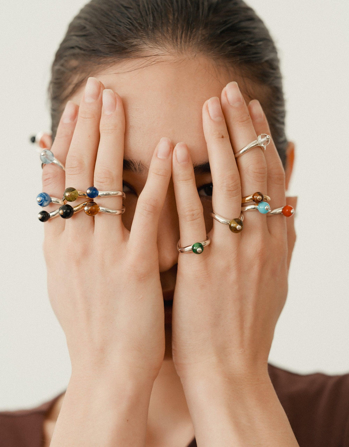 Hands covering face, showcasing multiple delicate rings with colorful stones on fingers, representing nature-inspired jewelry design. Pea Curve Ring | Large among the collection, embodying organic shapes and transformation. CLED