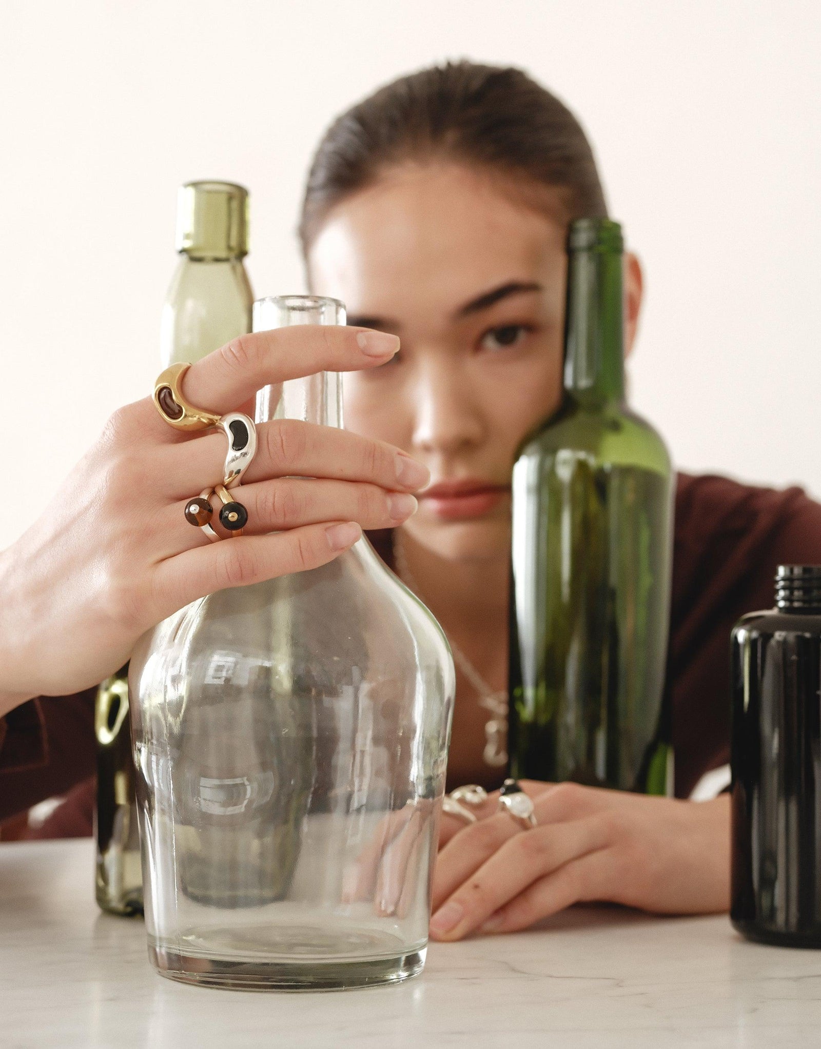 Person showcasing unique Bean Ring and other jewelry pieces, surrounded by glass and green bottles, demonstrating the natural-inspired design of the collection. Organic shapes reflect transformation and growth in legume-inspired jewelry. CLED Bean Ring