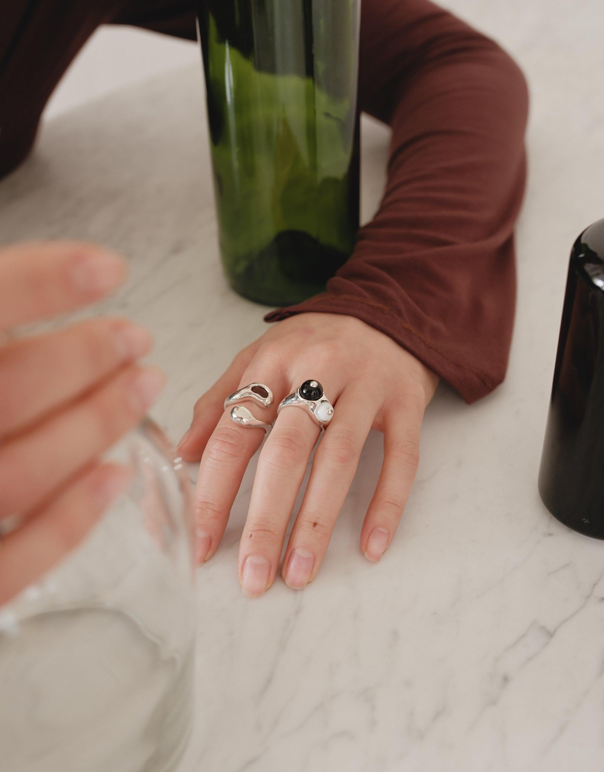 Elegant hand wearing the Pod Ring, showcasing its organic, nature-inspired design. The ring's unique shape reflects the beauty of legumes, set against a marble surface with green glass bottles, embodying growth and transformation. A perfect blend of natural elegance and modern style by CLED.