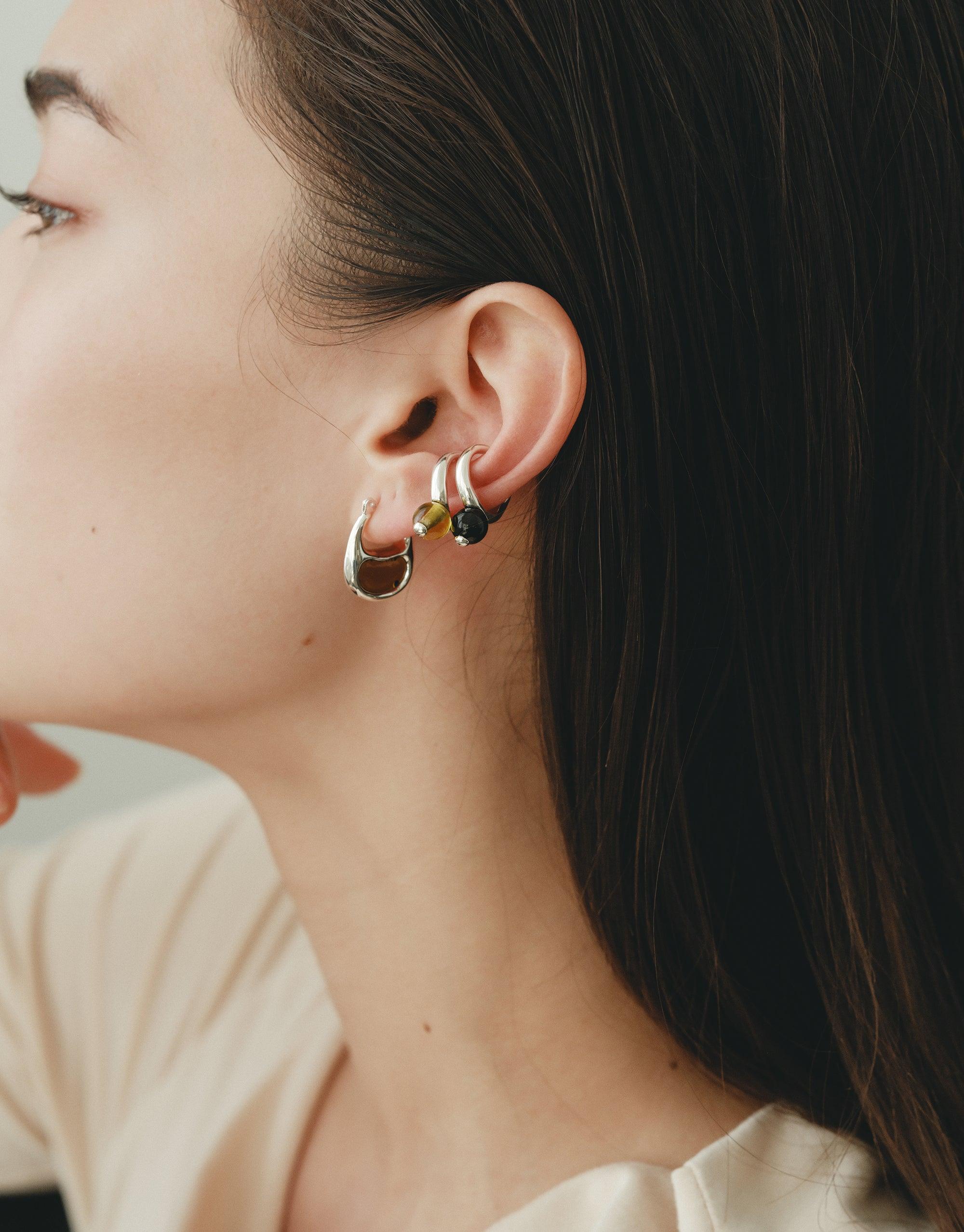 Close-up of a person's ear wearing small silver hoop earrings with a green bean-shaped charm, showcasing the delicate design of the Bean Hoop Earrings | Small. The earrings embody nature-inspired elegance and organic beauty. CLED