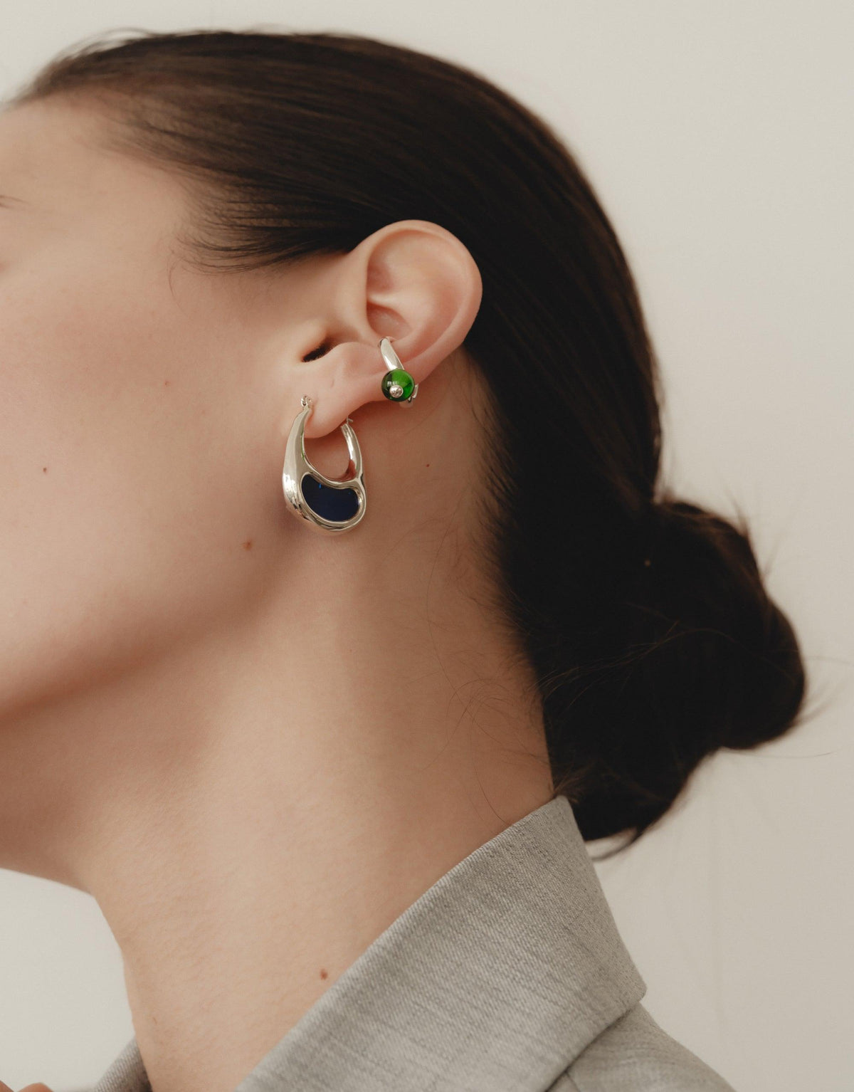 Close-up of a woman's ear wearing a small green pea-shaped ear cuff, showcasing the delicate and nature-inspired design. The ear cuff complements a larger curved silver earring, highlighting the organic shapes inspired by legumes. Pea Ear Cuff | Small by CLED