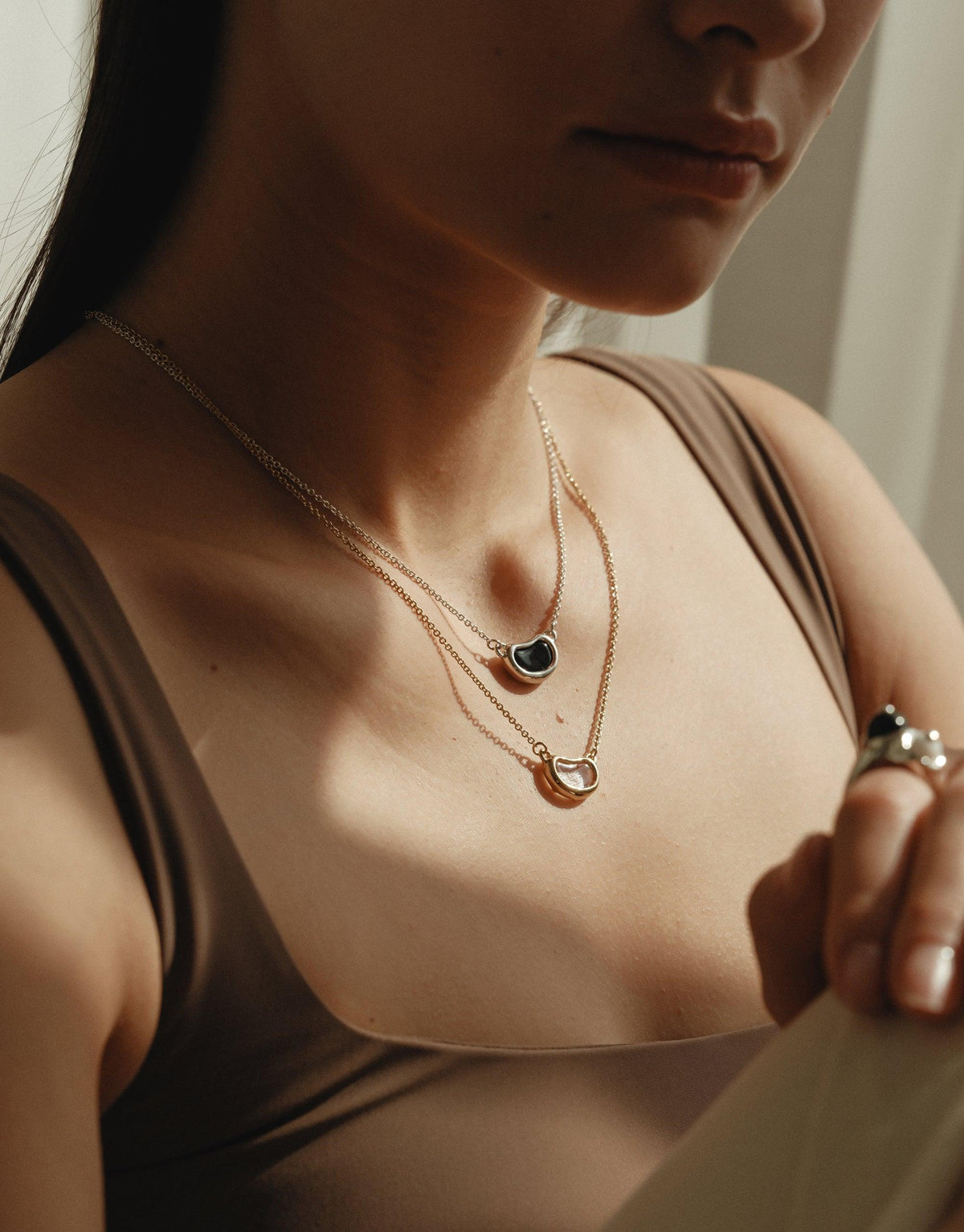 Close-up of a woman wearing layered delicate necklaces featuring organic bean-shaped pendants, showcasing the Bean Necklace's elegant design inspired by nature's curves and patterns. The necklace embodies growth and transformation, perfectly complementing the wearer's silhouette. CLED