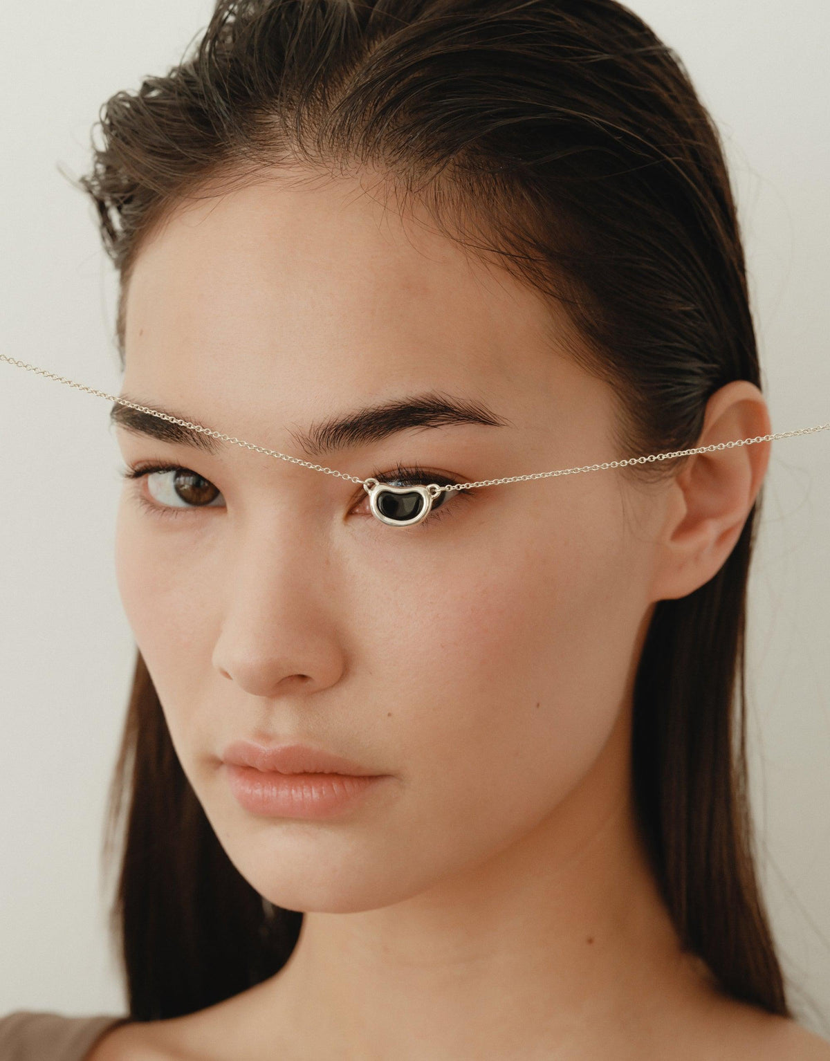 Close-up of a woman wearing a delicate silver chain necklace with a small bean-shaped pendant resting between her eyebrows, showcasing the unique Bean Necklace design inspired by nature's organic forms. CLED