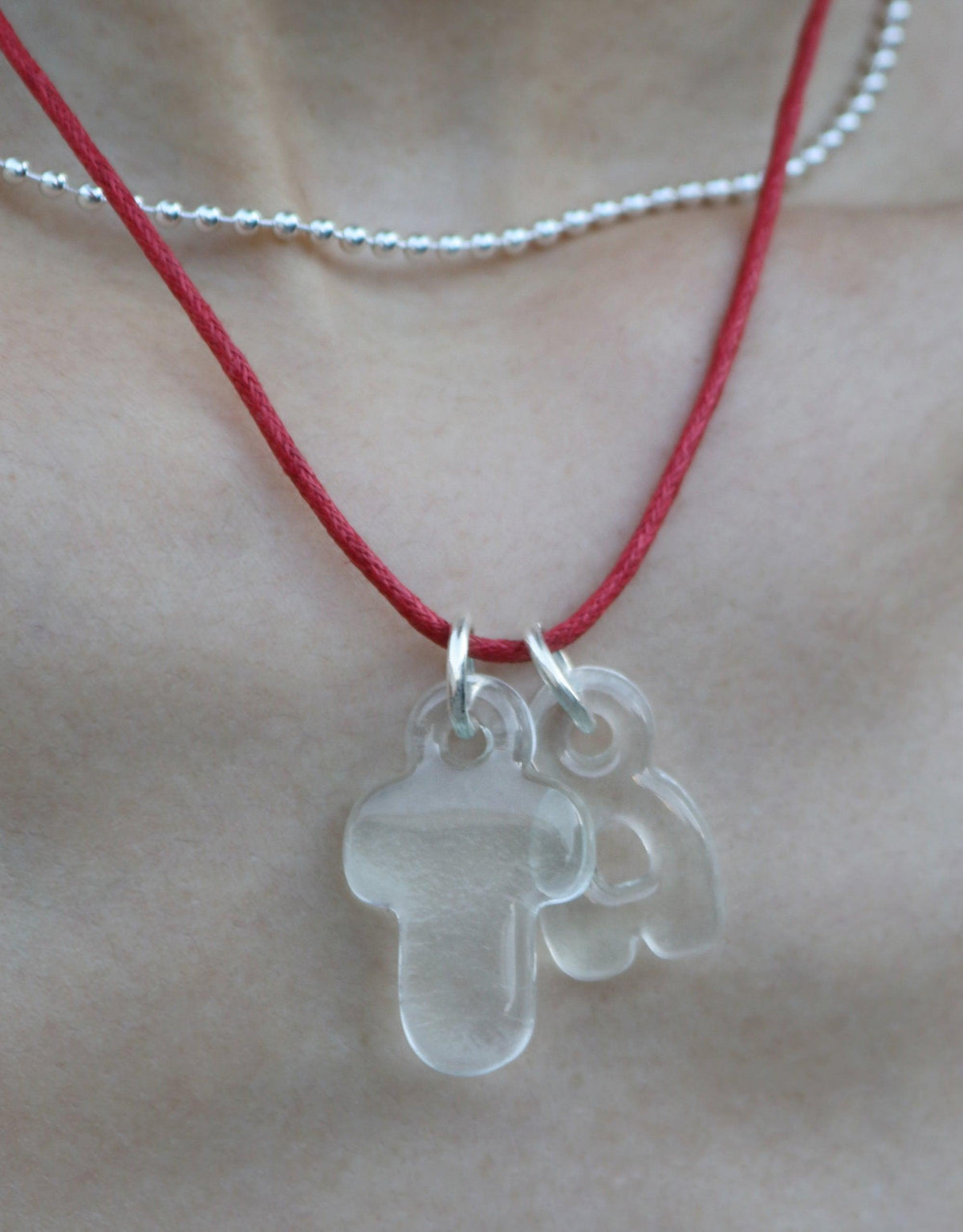 Eco-friendly bubble letter charms on red cord necklace, showcasing recycled glass alphabet pendants. Transparent, rounded letters for personalized expression. Layered with delicate silver chain. Sustainable jewelry by CLED. Letter Charm | Alphabet.