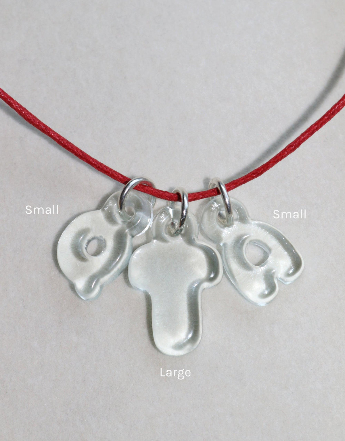 Adjustable red cotton cord necklace with three transparent charms: two small curved shapes and one large cross-like pendant. Versatile accessory for customizable styling. Cord Necklace by CLED.