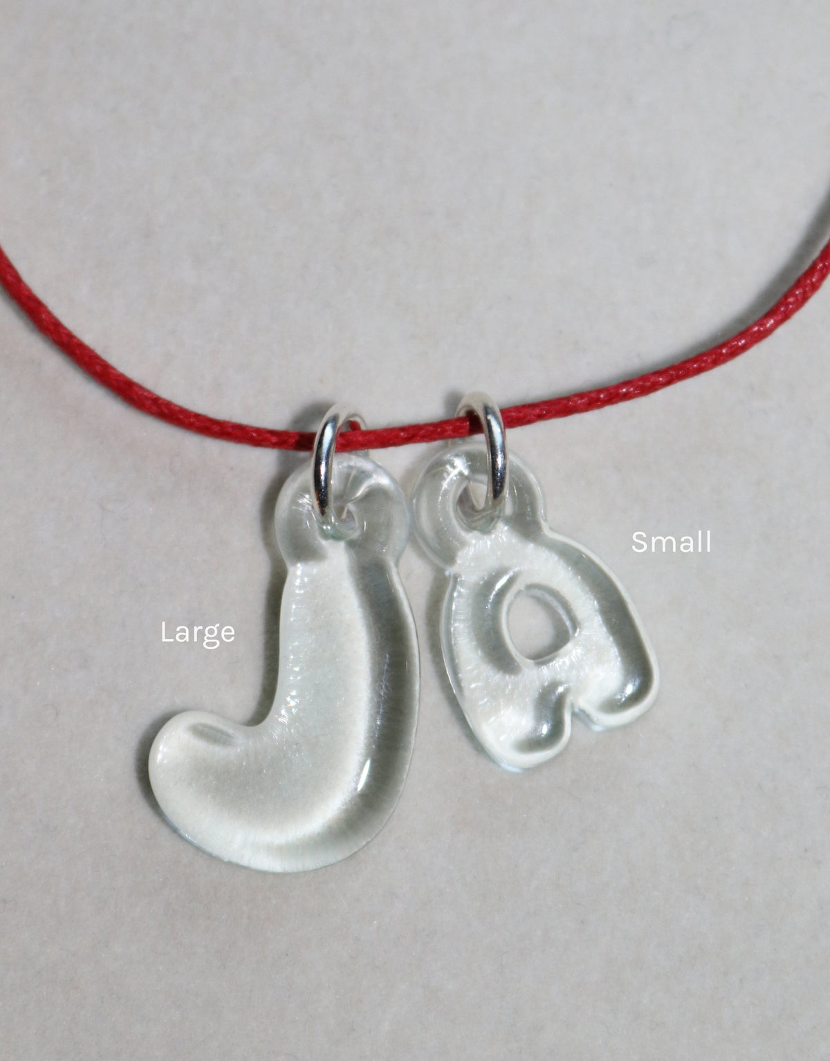 Eco-friendly bubble letter charms 'J' and 'a' in large and small sizes, made from recycled window glass, hanging on a red cord necklace. Playful and personalized jewelry for everyday wear. Letter Charm | Alphabet by CLED