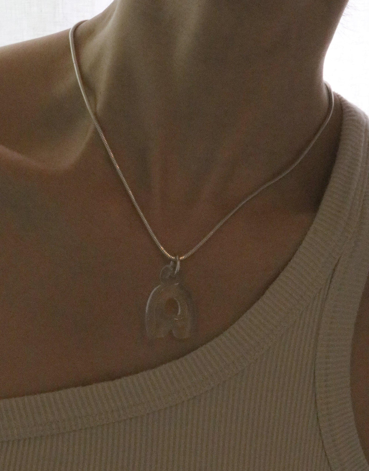 Transparent bubble letter 'R' charm necklace on silver chain, worn by person in beige top. Eco-friendly recycled glass pendant showcases personalized style. Letter Charm | Alphabet by CLED