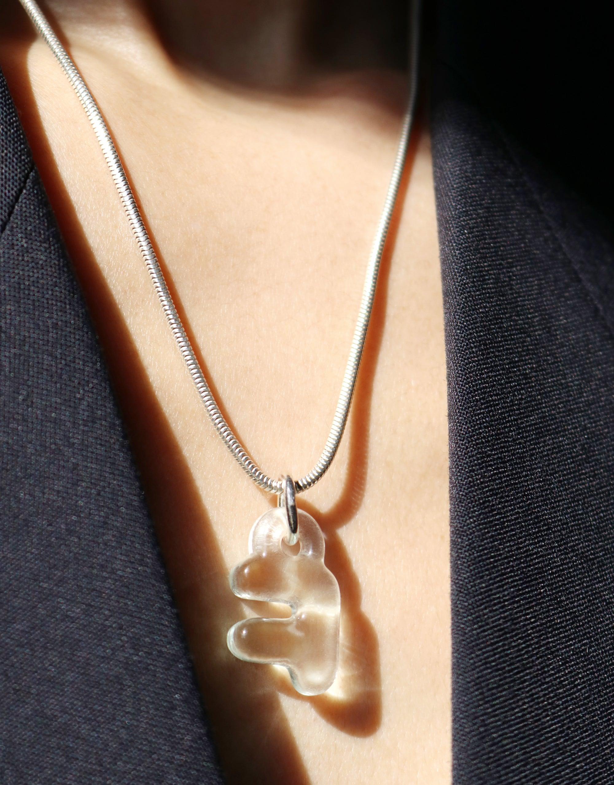 Close-up of a translucent bubble-shaped pendant on a silver chain necklace, worn against a contrasting dark and light background. The pendant appears to be a Korean Hangul character, showcasing the Letter Charm | 한글 Hangul product by CLED.