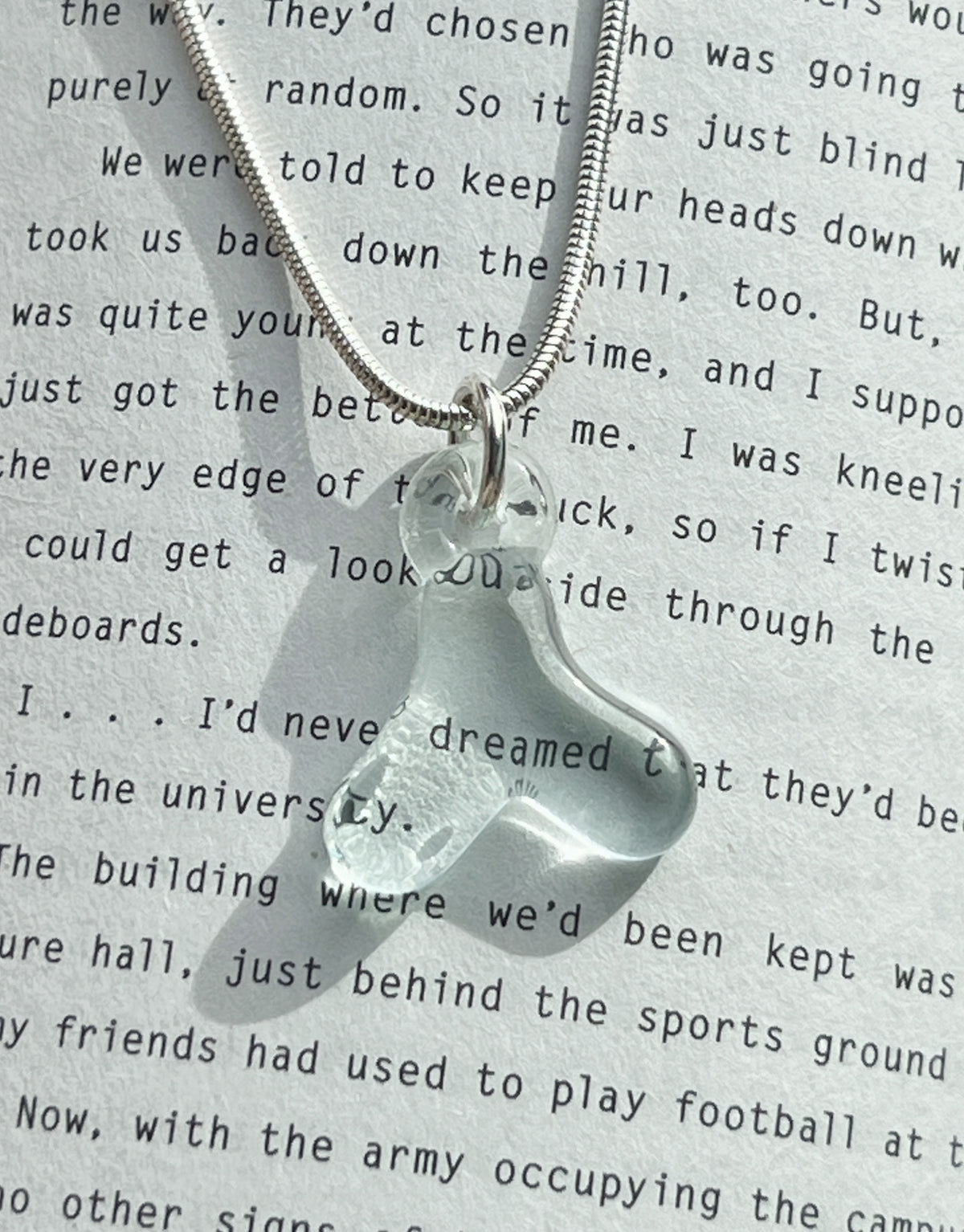 A snake chain necklace with a delicate glass Hangul letter pendant, set against a printed fiction background, highlighting the subtle beauty of the pendant