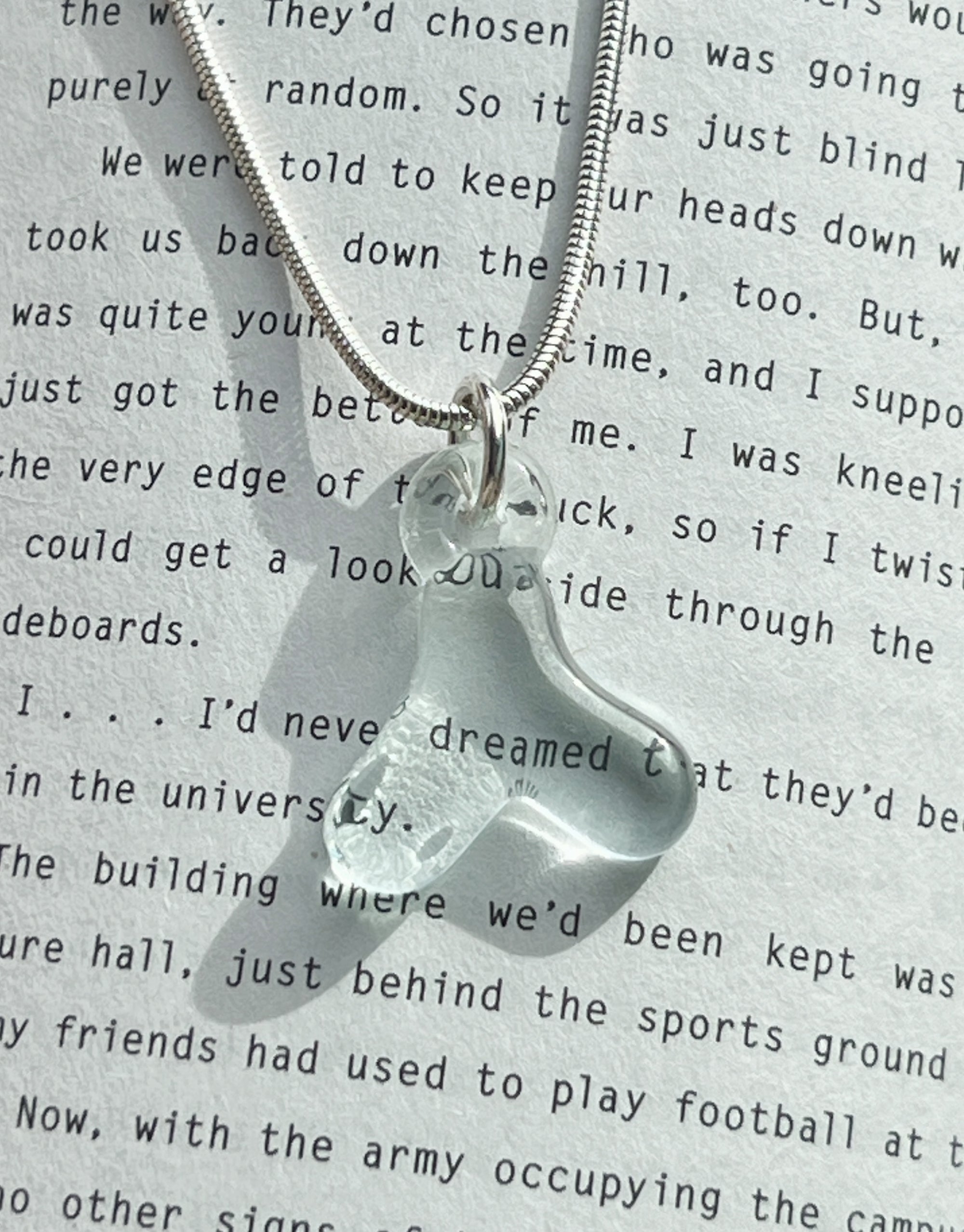 A snake chain necklace with a delicate glass Hangul letter pendant, set against a printed fiction background, highlighting the subtle beauty of the pendant
